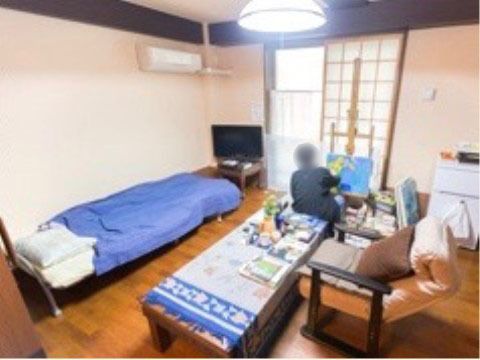 和の雰囲気を感じる居室。室内にはベッドやテーブル、椅子などの家具が置かれている。入居者様が絵を描いている。