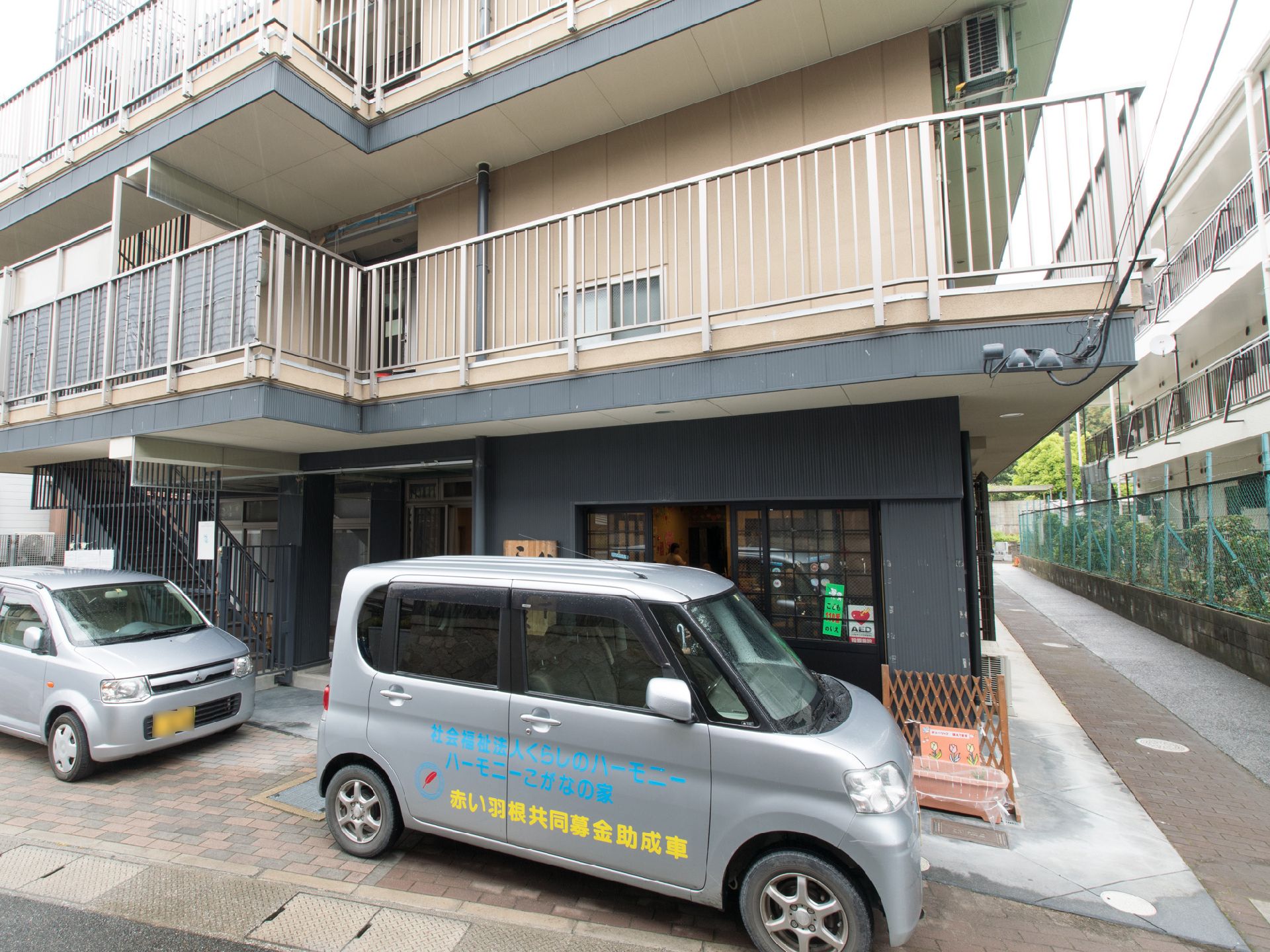 1階の玄関前には、黒い壁と引戸で統一されている。グレーの社有車が2台、施設の玄関前に縦列駐車されている。