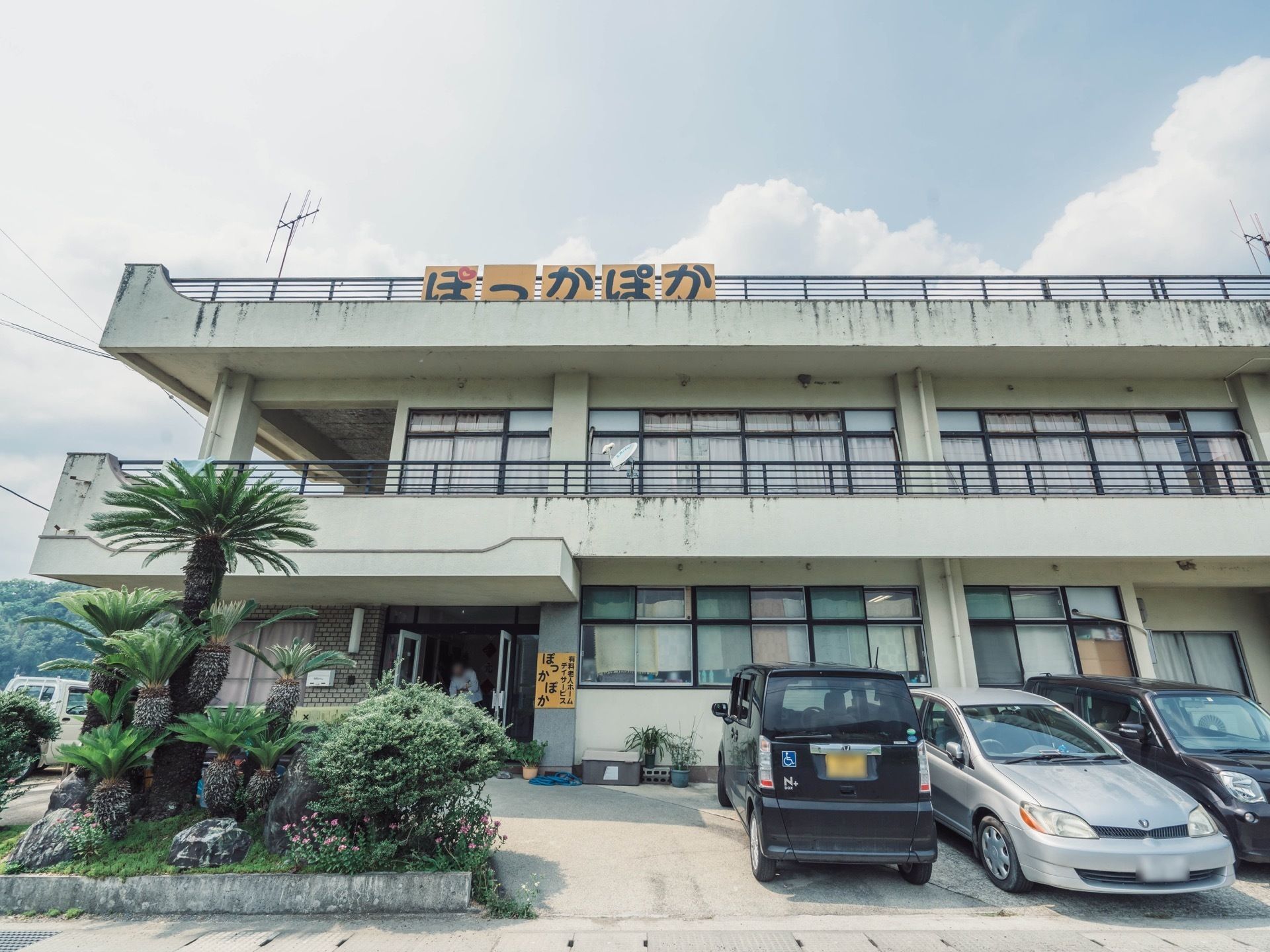 施設は2階建ての白い建物で、玄関の壁と屋上には施設名が書かれた看板が設置されている。建物の前には駐車場や植え込みがある。