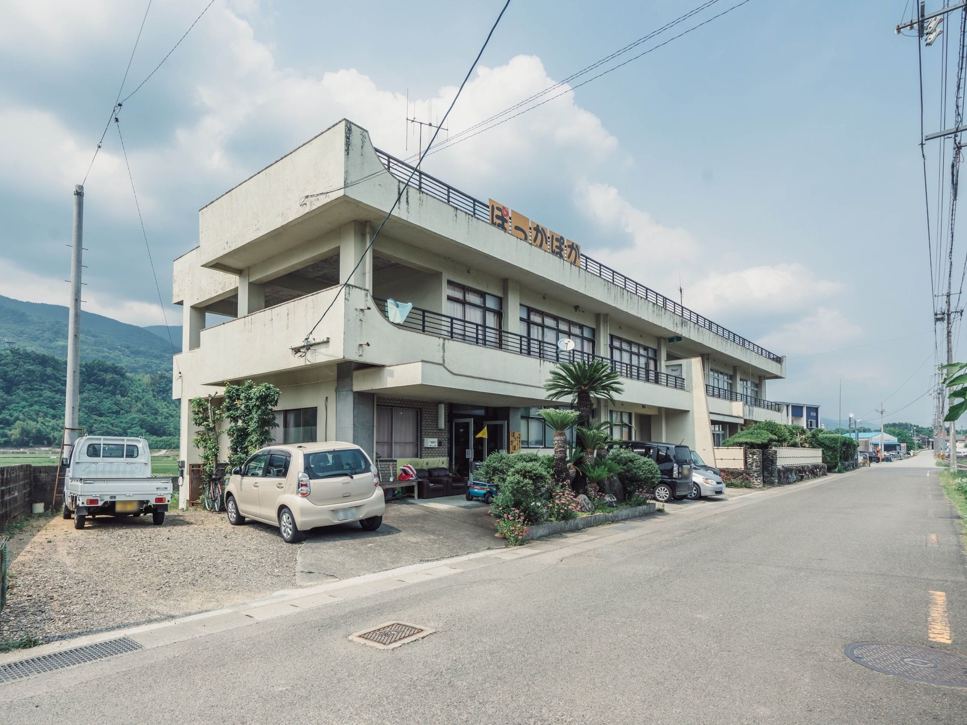 施設は白い2階建ての建物になっていて、2階と屋上には手すりが設置されている。建物の左側と中央付近には車が停められている。