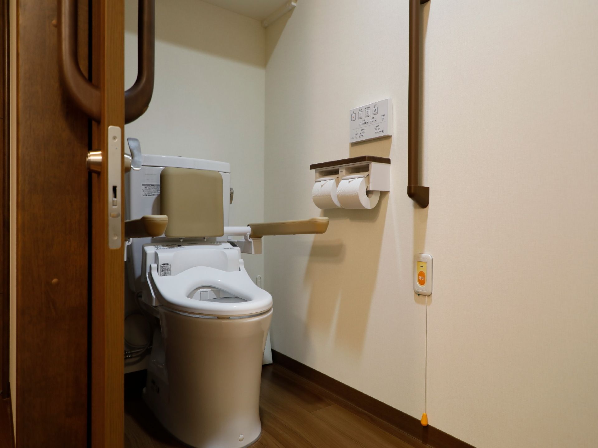 トイレは木目の引き戸で、開閉が安全にできる。壁に手すりが付いており、便器にも可動式の背もたれと手すりが設置されているので安心である。