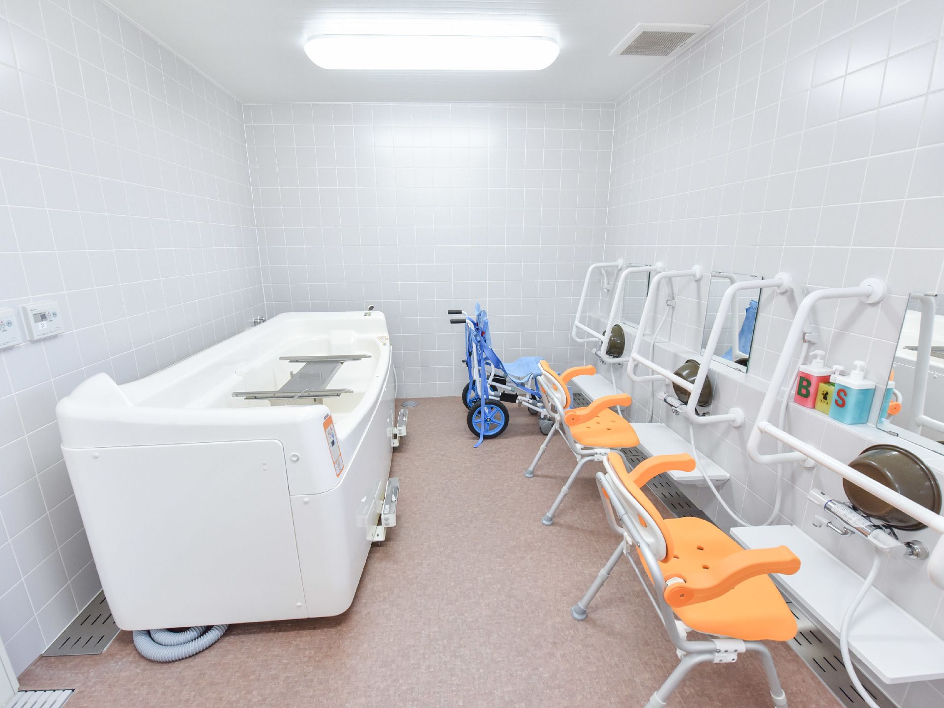 浴室の洗い場には、青い補助椅子とオレンジ色の補助椅子が2つ並べられている。白い大きな浴槽も設置されている。