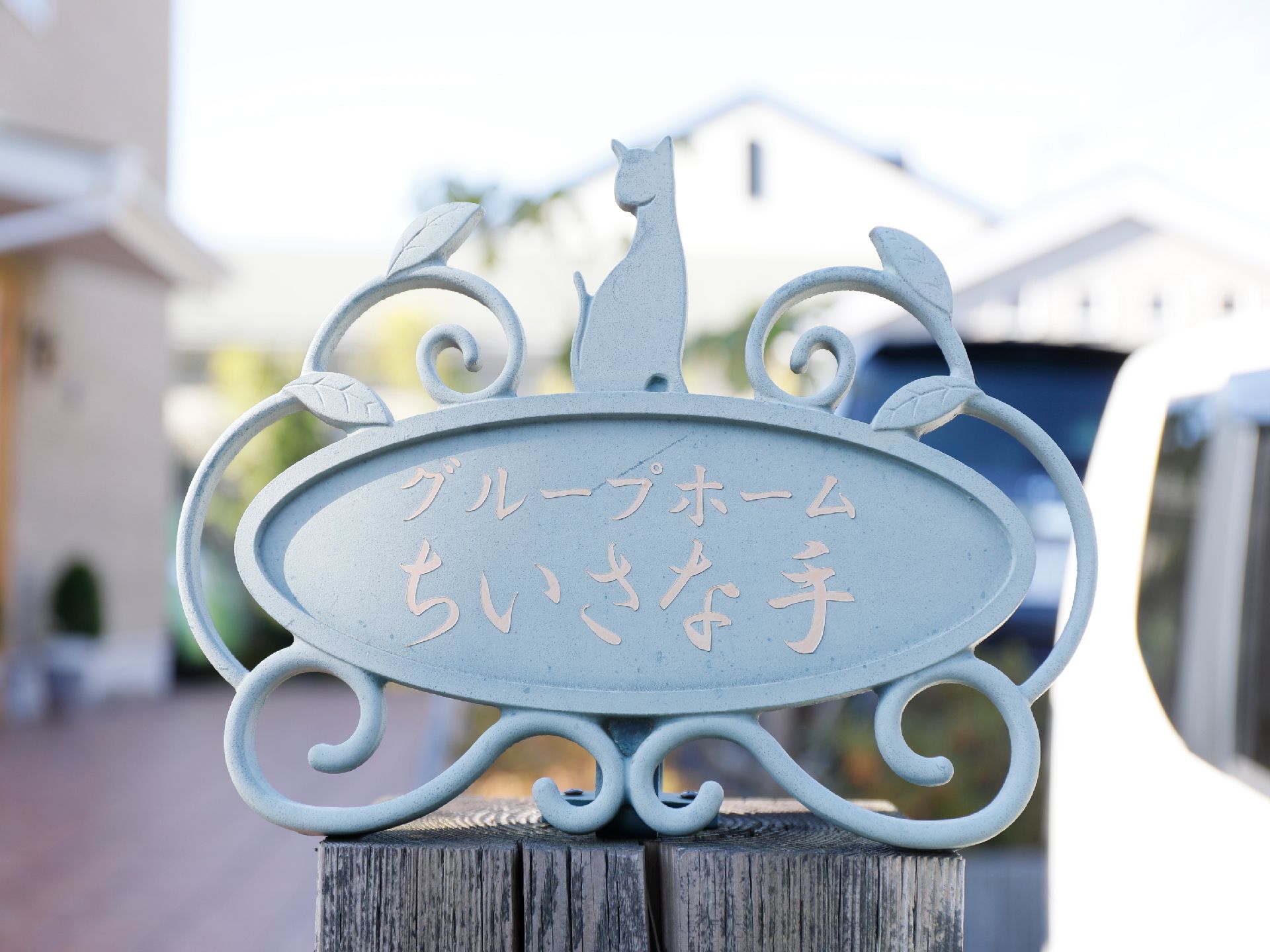 木材の上に、看板が置かれている。ネコと植物の装飾がほどこされた楕円形の看板に、施設名が表記されている。