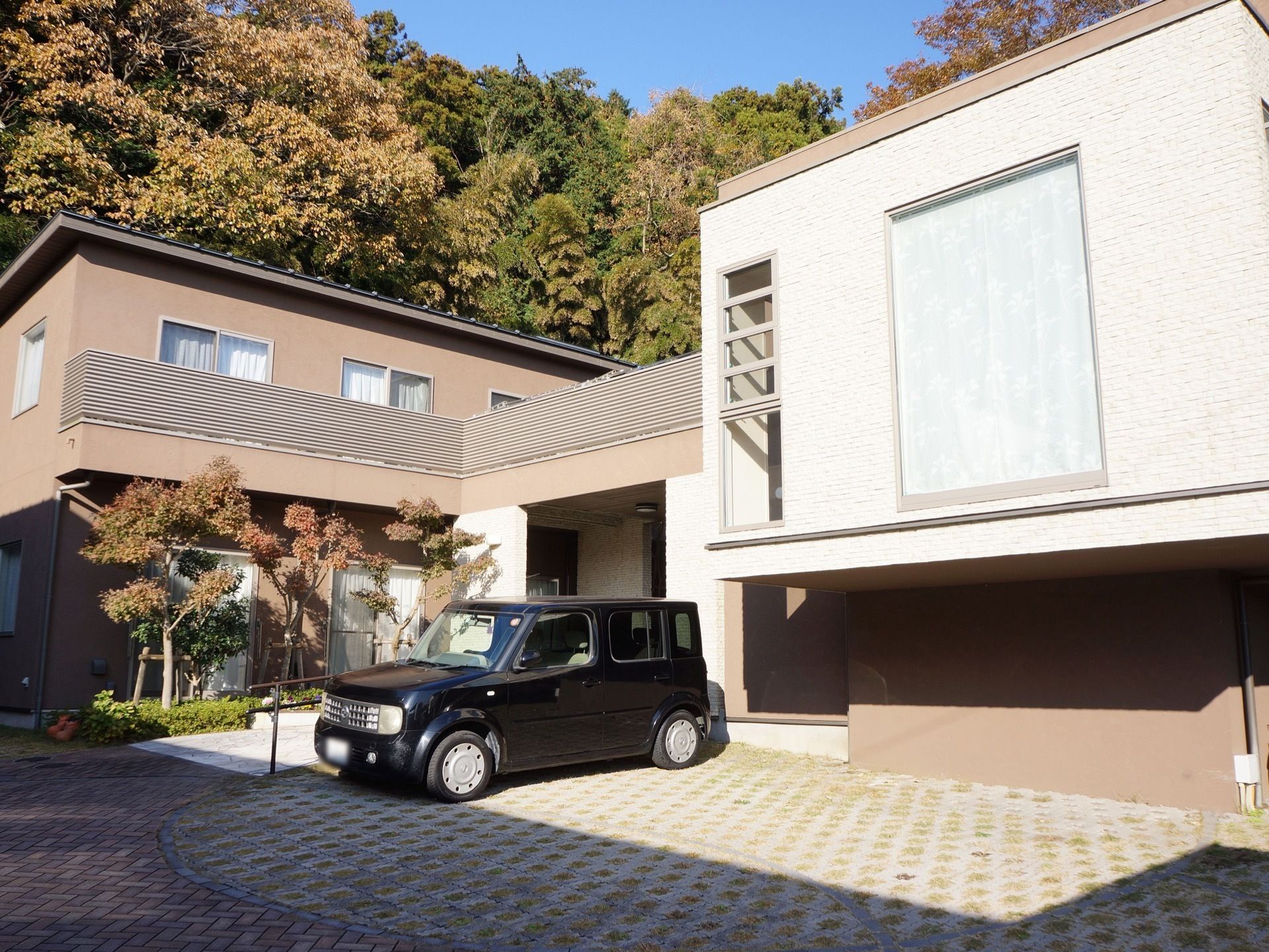 一軒家風の建物が2つあり、つながっている。建物前にバリアフリーの駐車スペースがあり、黒い車が1台駐車している。