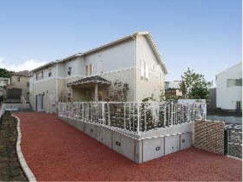 緩やかな坂になった赤と茶色の通路が続いた先に、二階建ての白い外壁の建物がある。庭は白色のフェンスで囲われている。