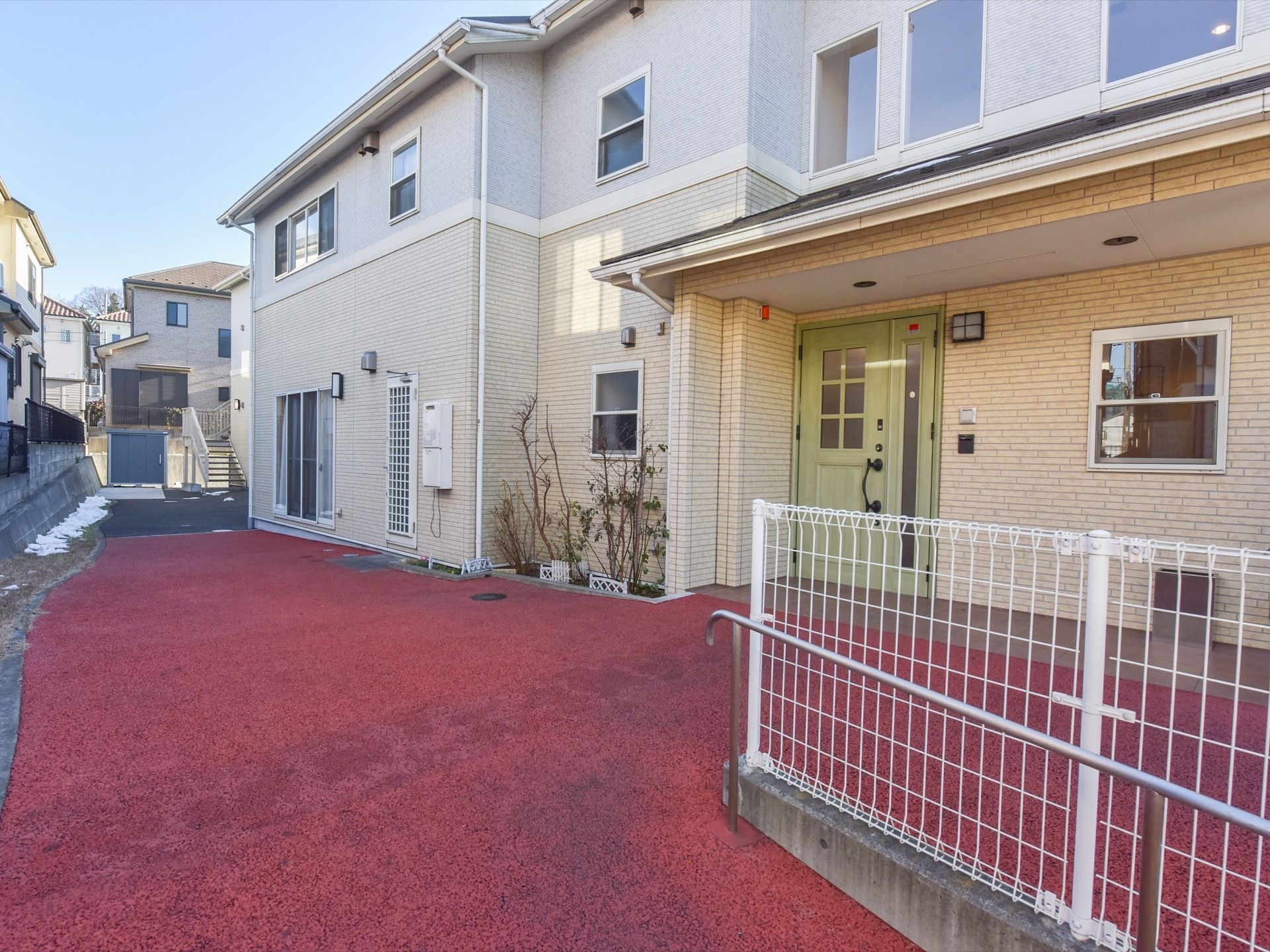 一軒屋風の二階建て建物が、右手に建っている。建物前の地面は赤く、低木が植えられた小さなスペースがある。手前に手すりが設置されている。