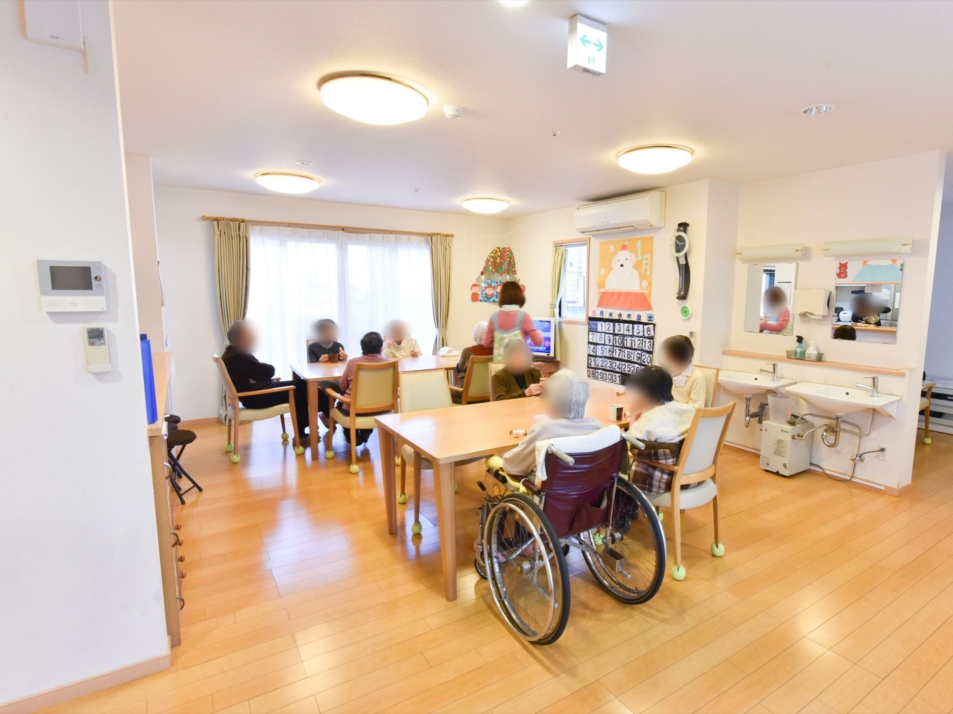 リビングにテーブルセットが２つあり、スタッフと入居者様たちが集まっている。車椅子の方もいる。椅子の脚には、滑り止めが付いている。