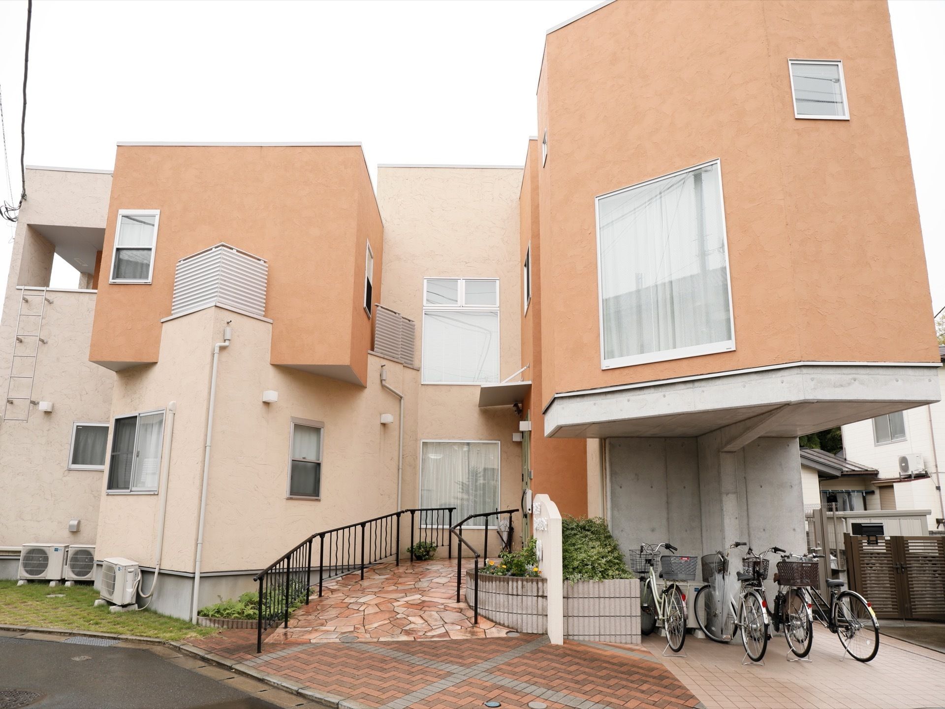 モダンでおしゃれな外観の２階建て施設には、建物前に手すり付きのスロープや駐輪場が設けられている。花壇が設置されている。