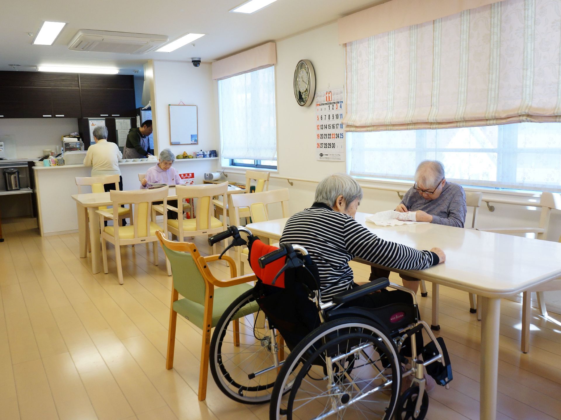 ダイニングの床は、木製の板が組まれたフローリングである。室内では椅子に座った入居者様に加え、車椅子の入居者様もいる。