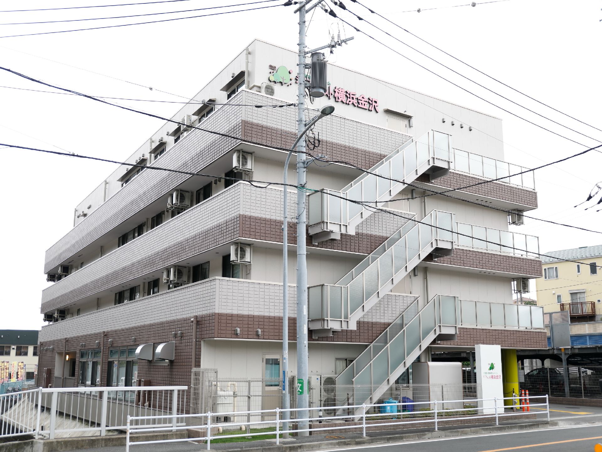 施設全体が見えるように撮影した写真で、施設は、三階建てで、メッシュフェンスで敷地は囲んである。外階段があり屋上まで登っていくことができる。