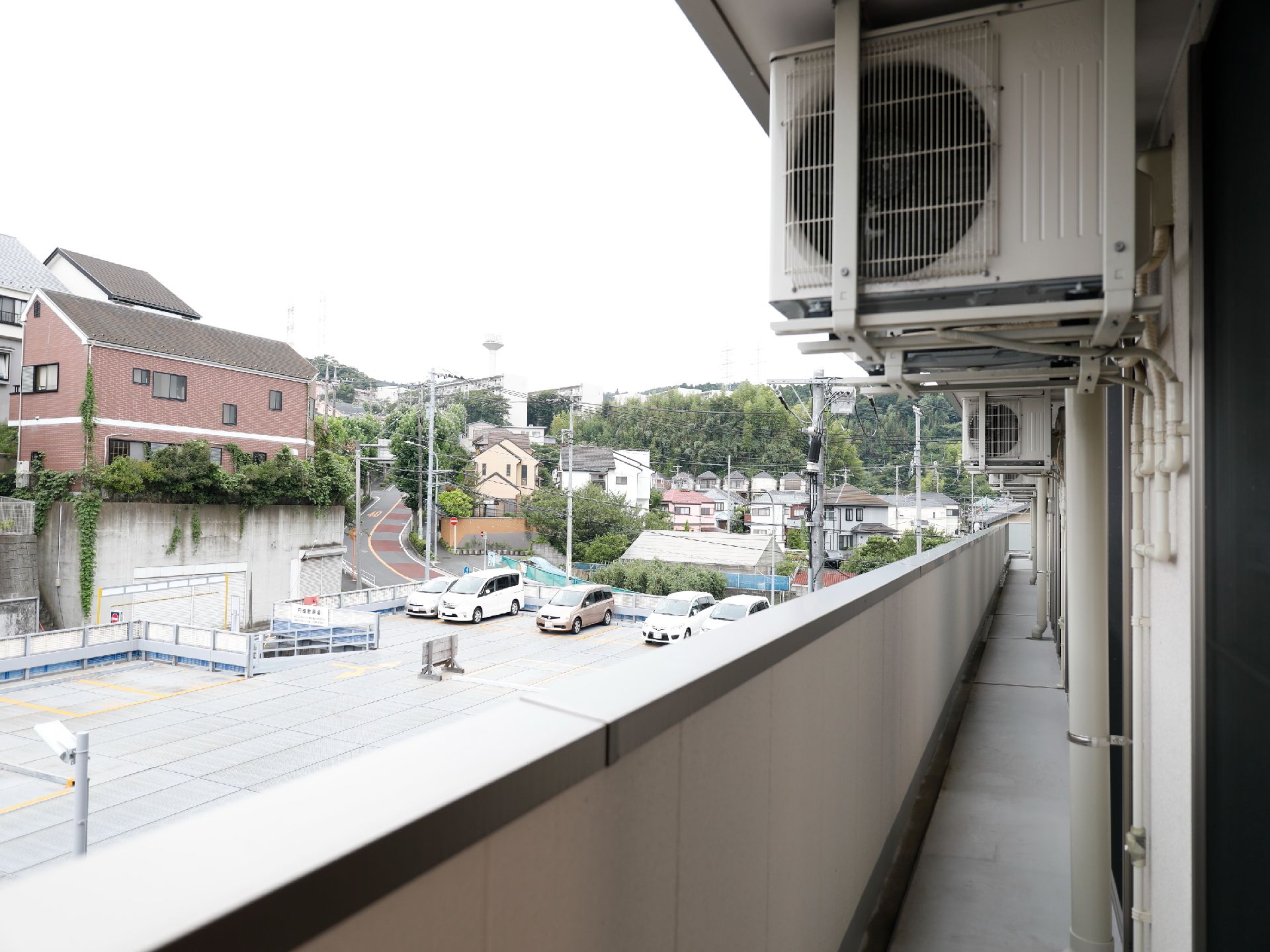 施設のベランダから見える風景。階下には、駐車場がある。坂のある街で、住宅が林立しているのが見える。街路樹がある。