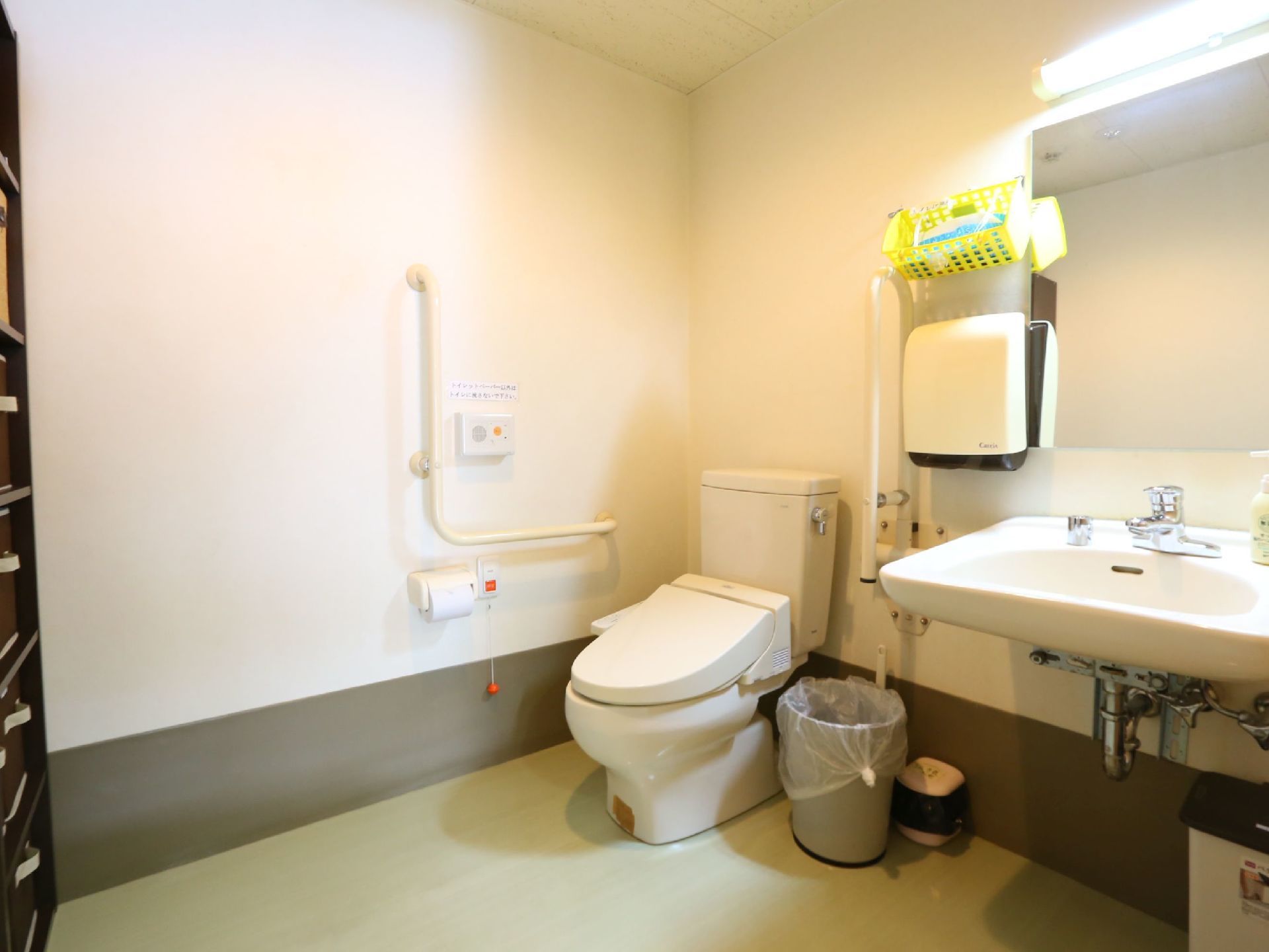 トイレには温水洗浄便座が設置されており、壁に操作パネルが取りつけられている。手すりの下にトイレットペーパーと非常ボタンがある。