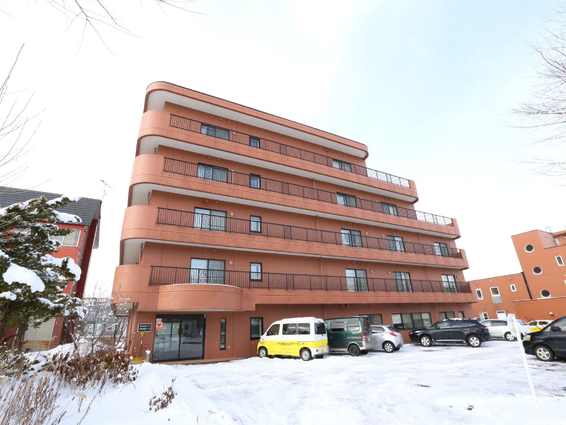 茶色の外壁の５階建ての施設である。ベランダは黒い柵になっている。施設の前は駐車場があり、たくさんの車が停まっている。