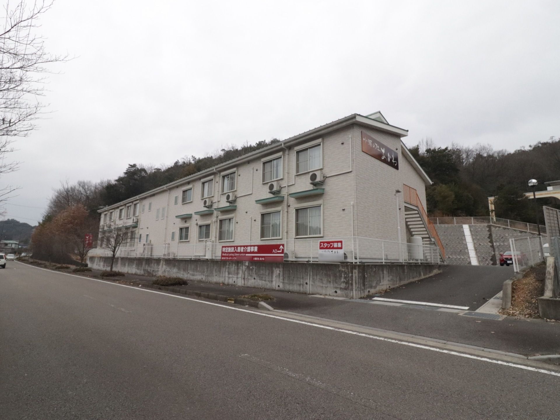 ベージュ系を基調とした外壁に施設名のプレートが設置されている。２階建て。周辺は大きな建物がなく木々に覆われている。