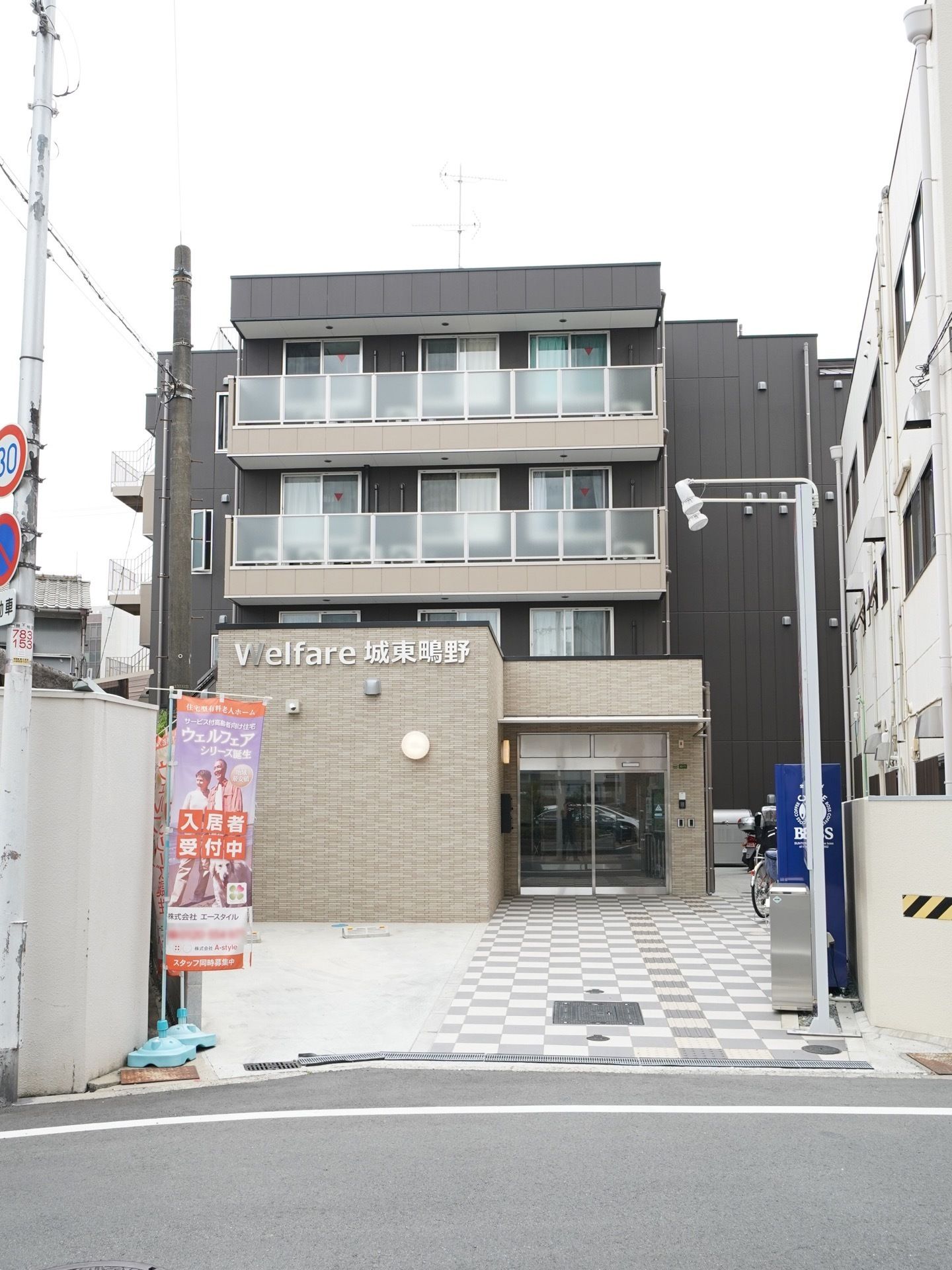 地上４階建てのマンションのようなグループホームである。道路沿いに旗が立てられており、自動販売機も設置されている。
