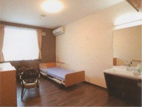 居室には大きな窓があり、エアコン、ベッド、洗面台が設置されている。フローリングの床の上にはテーブルと椅子が置かれている。