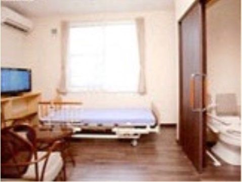 居室には引き戸で区切られたトイレがあり、大きな窓が設置されている。フローリングの床にはベッド、テーブル、椅子、棚が置かれている。