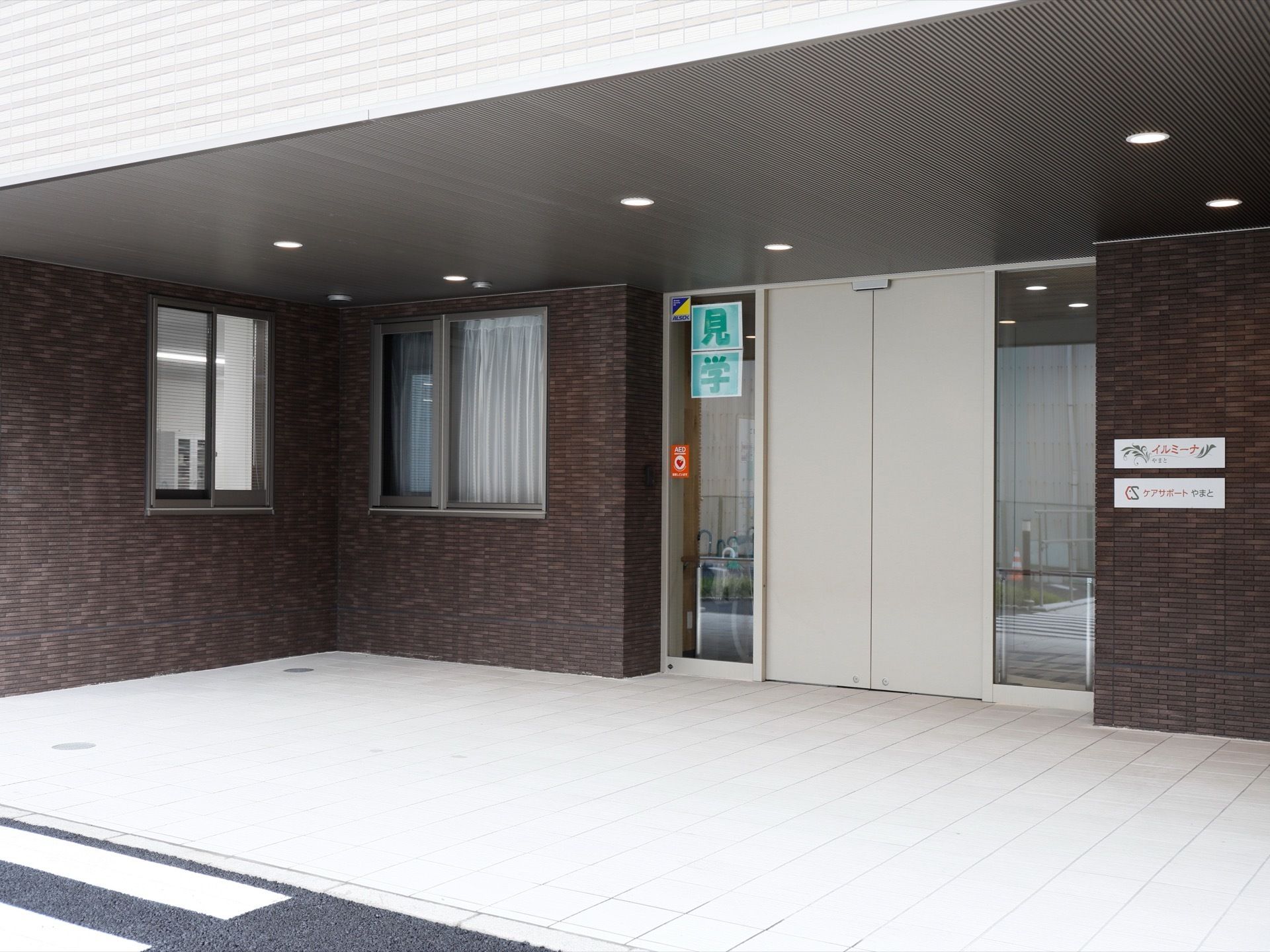 施設の玄関はグレーの自動ドアで、両脇はガラス張りにしている。右側の壁には施設名とロゴマークを入れたプレートを設置している。