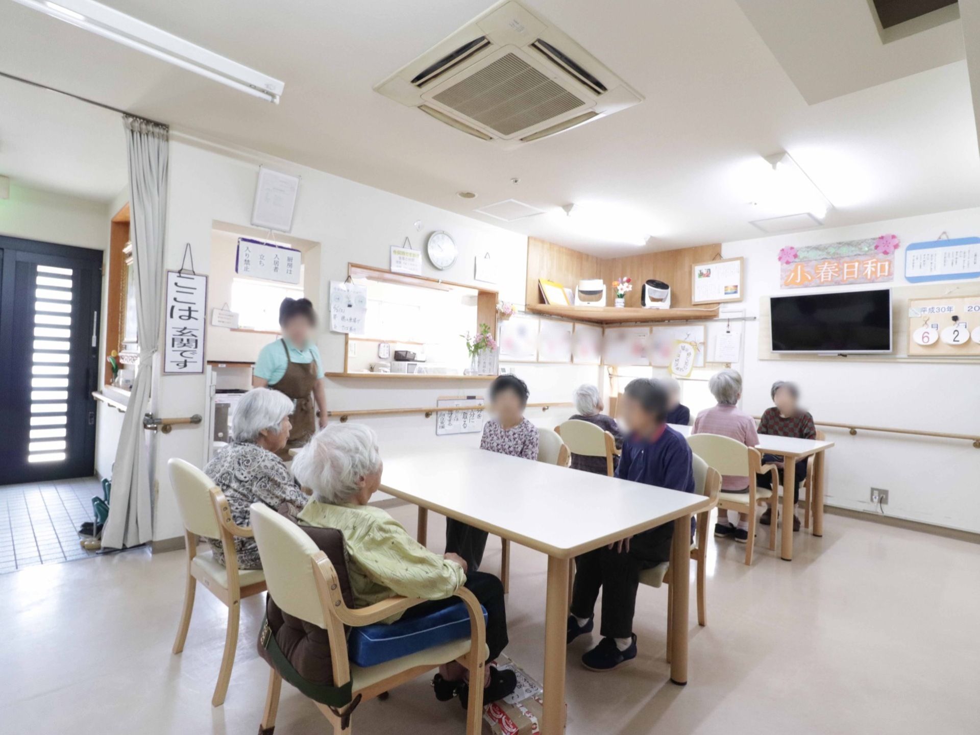入居者様が、テーブルを囲んで椅子に座っている。壁には、大型テレビが設置され、掲示物も貼られている。玄関を示すプレートがある。