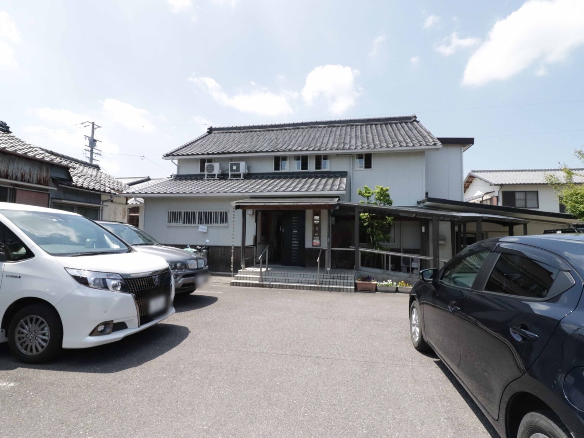 瓦屋根に白い壁の建物である。入口には、階段とスロープがついている。建物の前は駐車場になっており、車が３台停まっている。