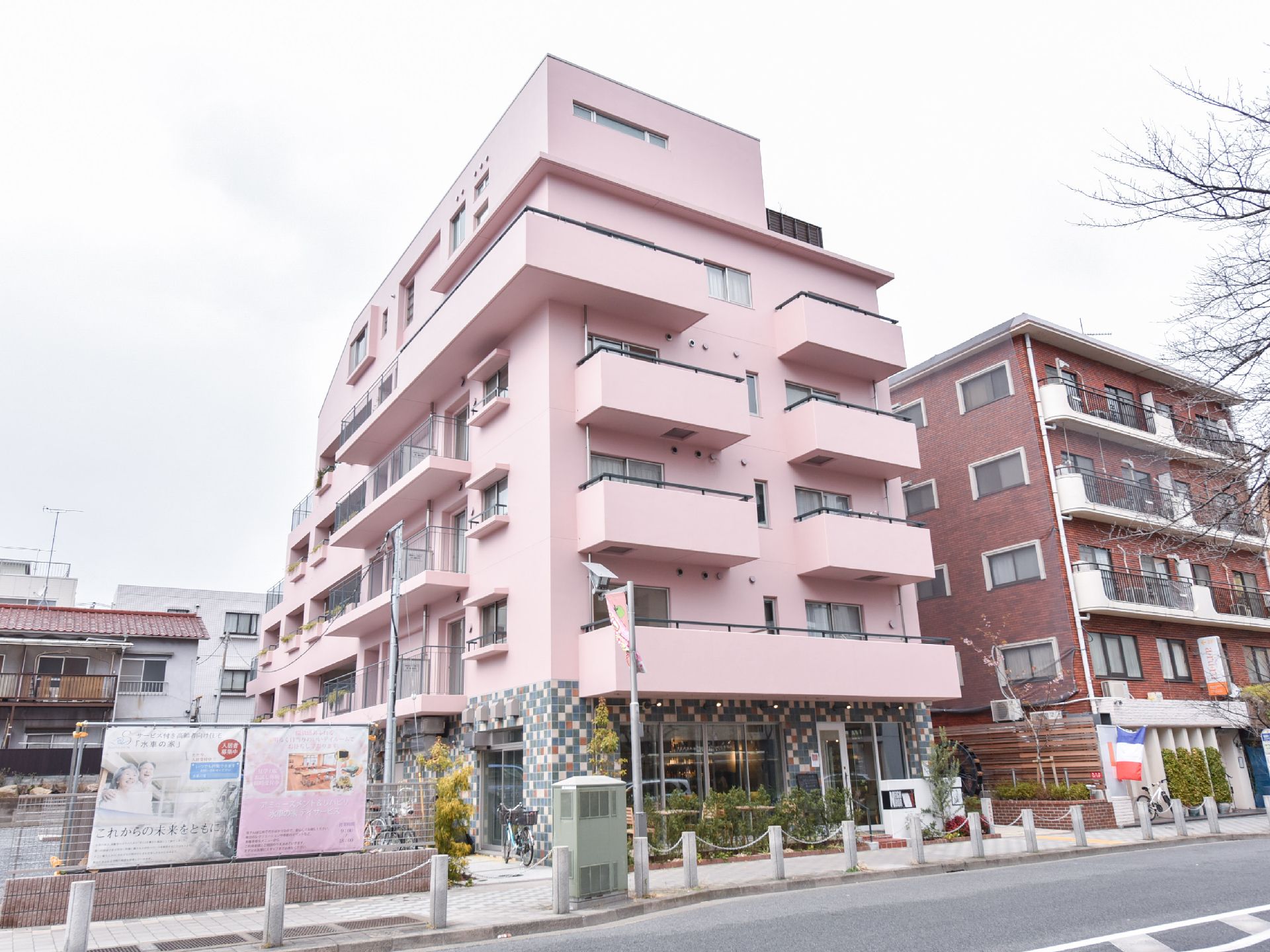 建物全体がピンク色である。ストライプのフェンスと目隠しフェンスのあるベランダがある。一階部分の外壁はタイルである。