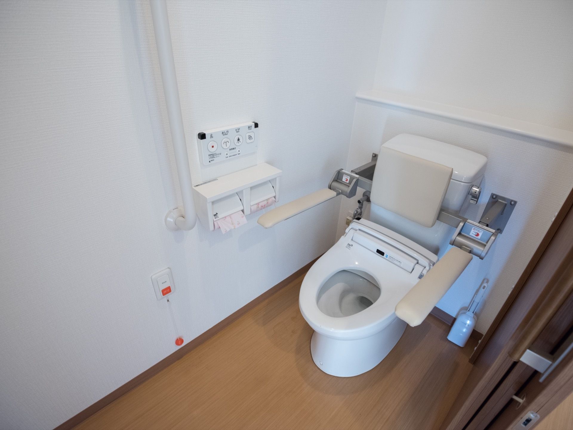 トイレの便座の両側に、可動式の手すりが設置されている。壁には縦方向の大きな手すりもあり、その下に呼び出しボタンがある。