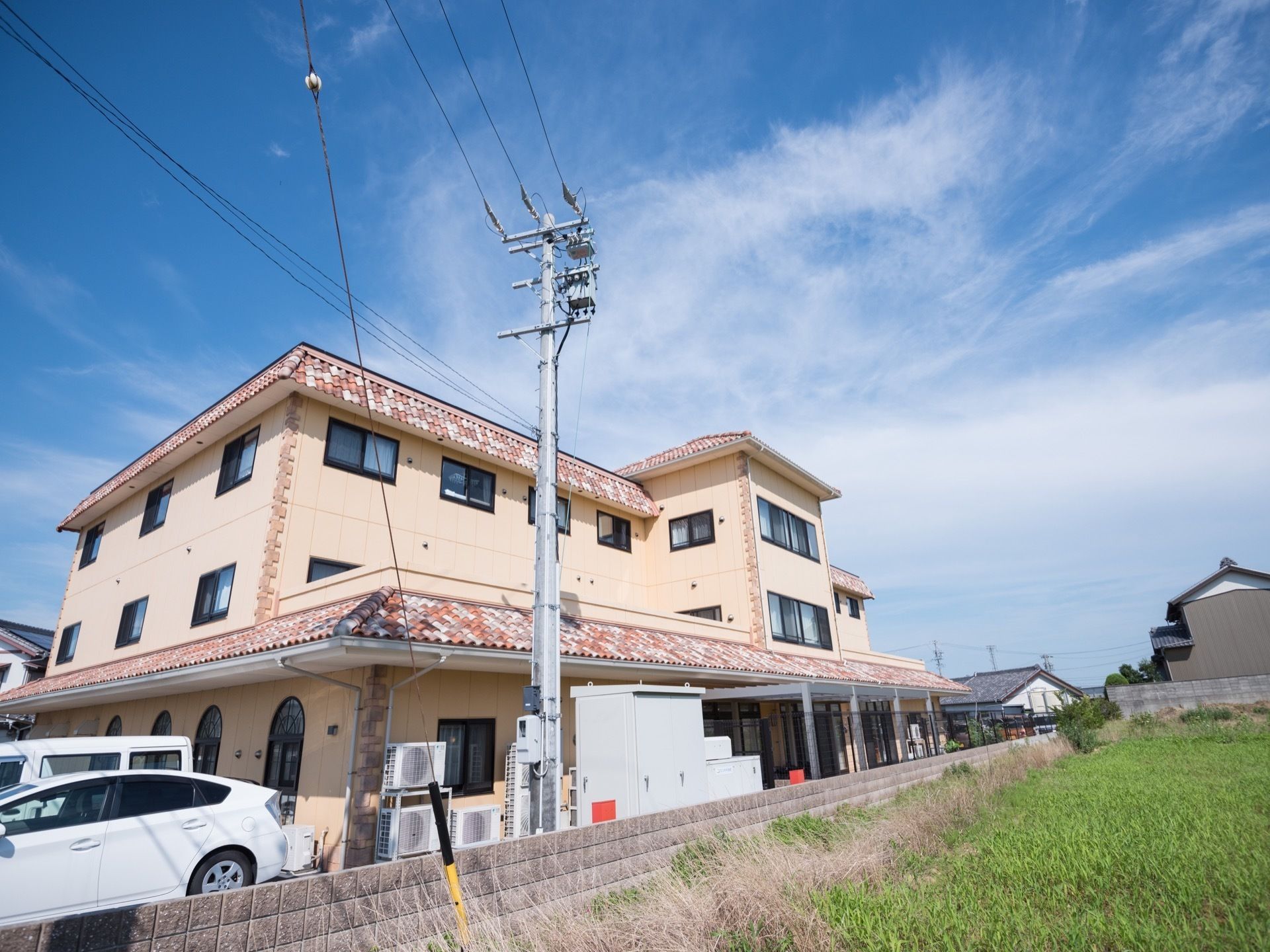 薄い黄色の外壁と、色違いの瓦の屋根がある３階建ての施設である。隣の敷地には建物がなく、一面に草が生えている。