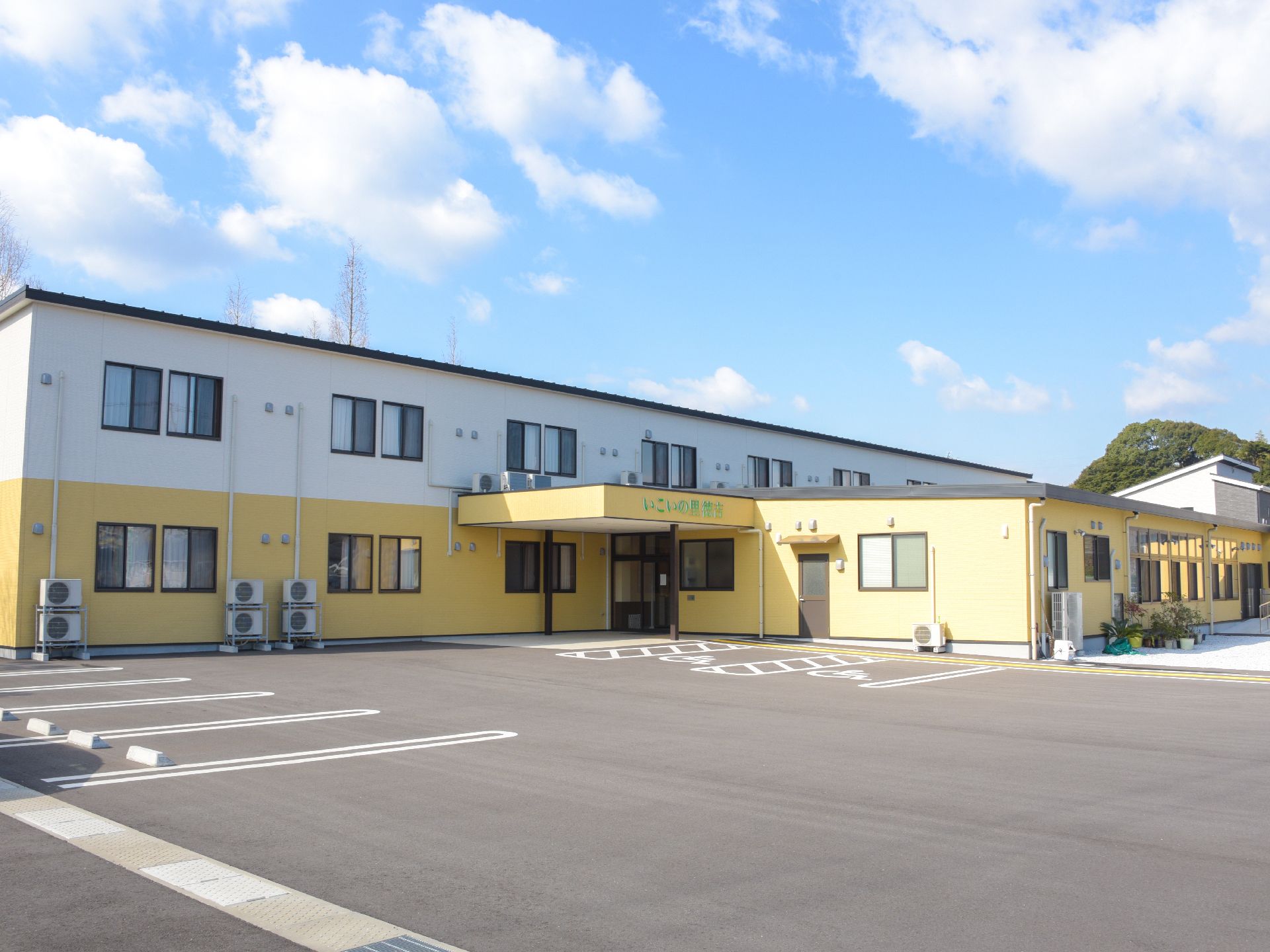 施設建物は2階建てになっている。外壁は1階が黄色く、2階は白で分けられている。車の駐車スペースが広く取られている。