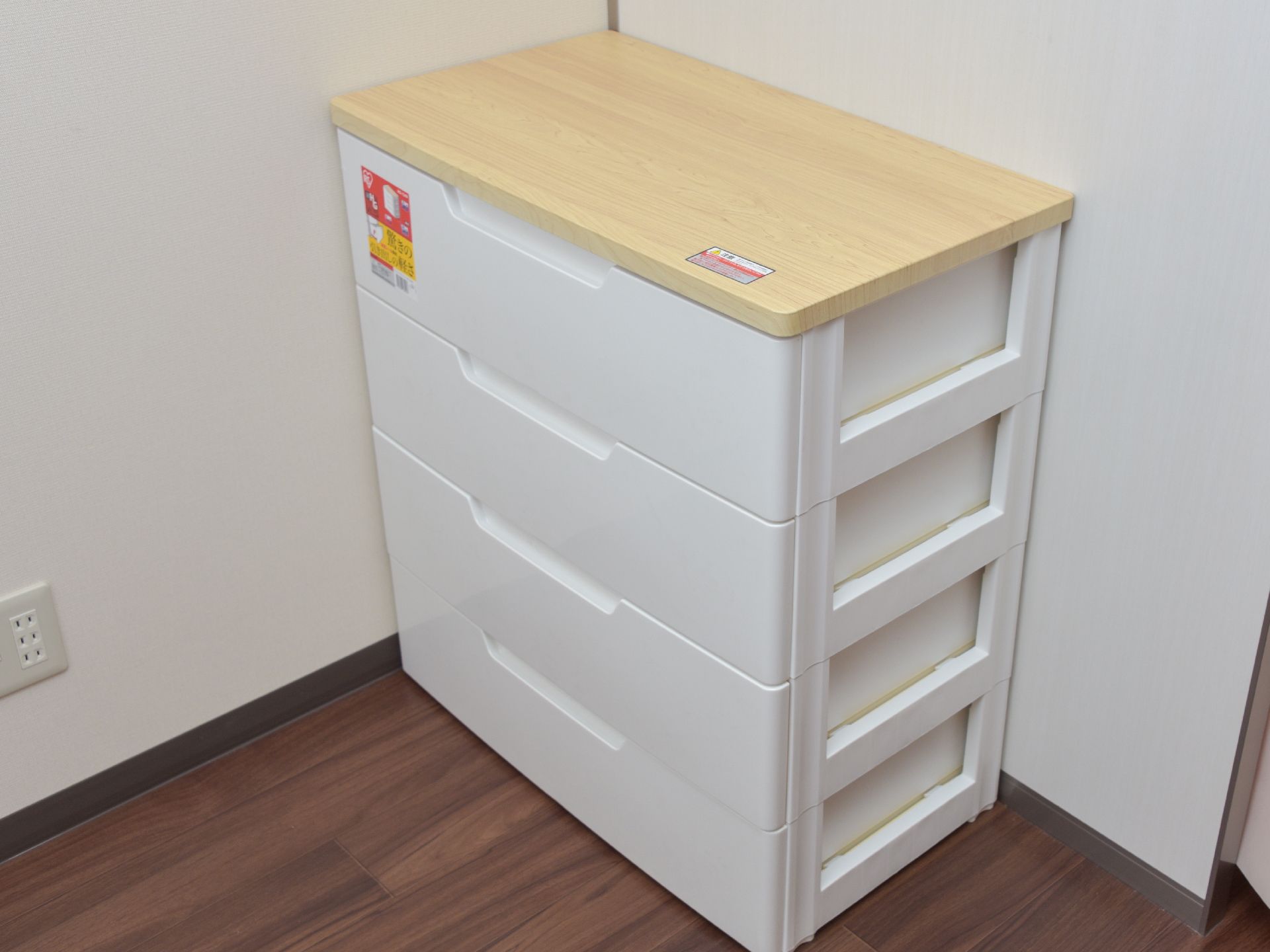 部屋の一角には白とナチュラルな木目調の4段ボックスが置かれている。すぐ横の壁にはコンセントが3つさせるようになっている。