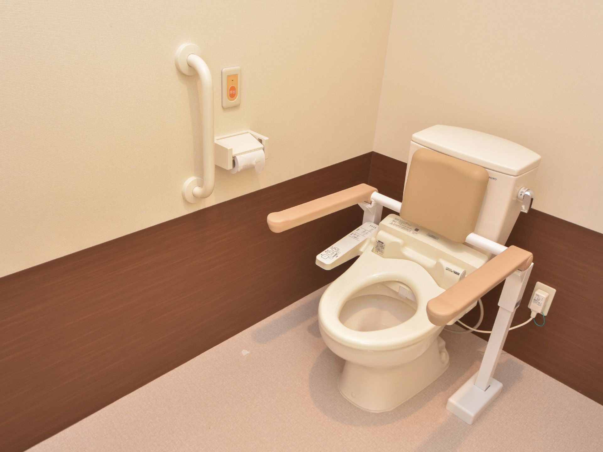 トイレの左右には手すりがあり、ベージュ色の背もたれも設置されている。左側の壁にも、白い頑丈な手すりが付いている。