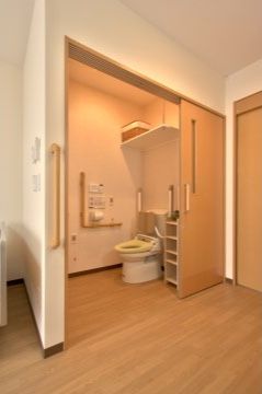 居室の入口近くに設置されたトイレである。便器横に収納棚が置かれており、壁にはＬ字型の手すりが設置されている。