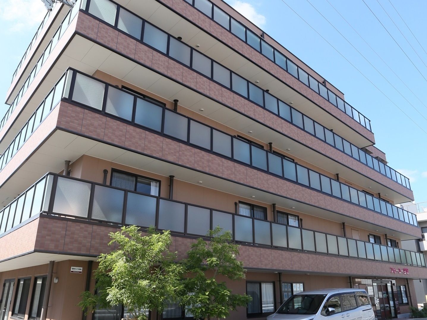 施設は５階建てのモダンな建物である。建物の前が駐車場となっており、数台が停まっている。駐車場の横に庭木が植えられている。