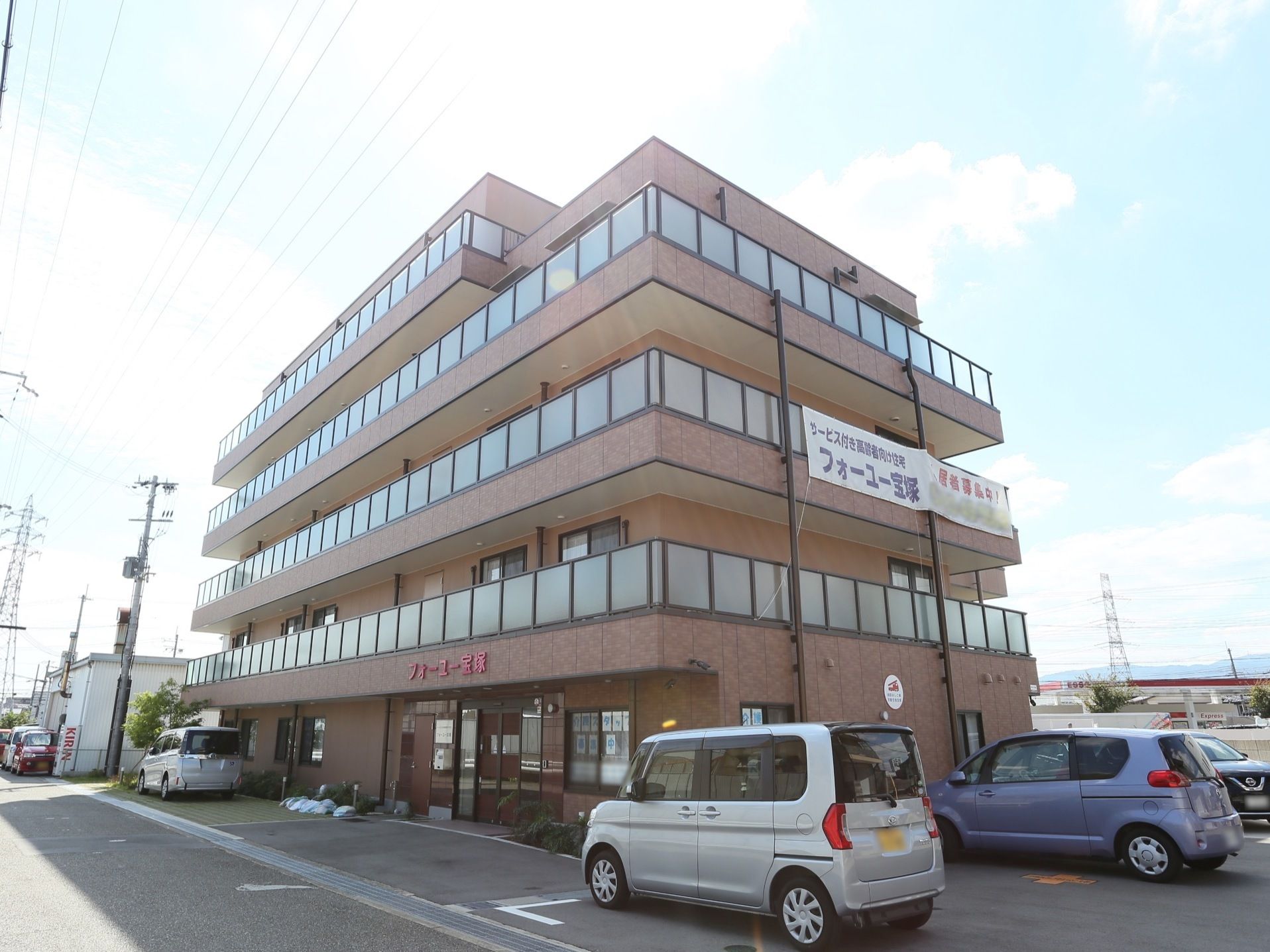 施設は茶色の５階建てである。施設の横に駐車場が設置され、数台が駐車している。玄関上の外壁に施設名称が入っている。