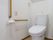 白壁で清潔感のあるトイレである。洗浄機能付きの便座で、横の壁には、縦、横の手すりが設置されている。隣には緊急通報ボタンが取りつけられている。