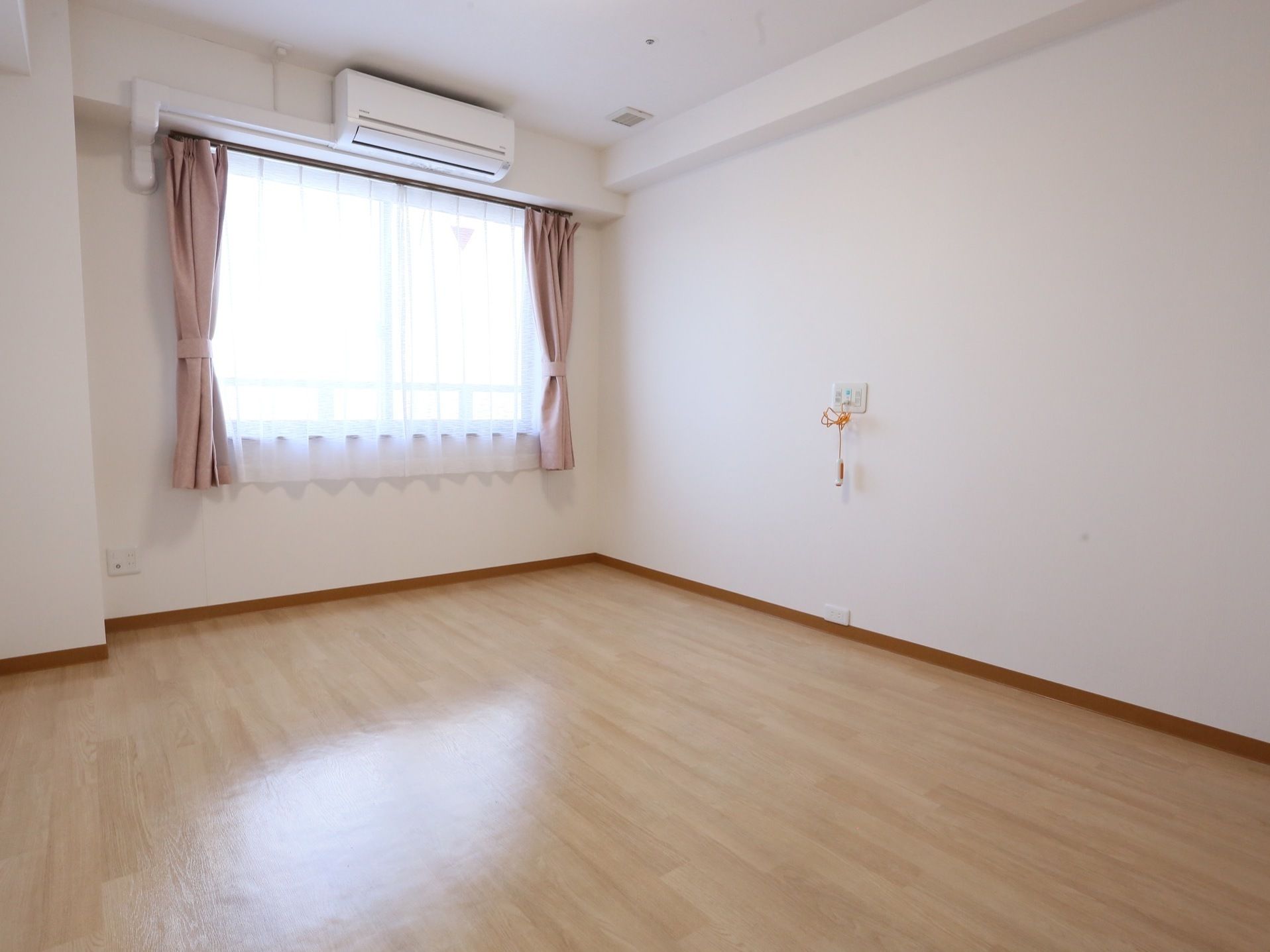 大きな窓から光が差し込む明るい部屋である。窓にはカーテンが設置されている。窓上の壁にエアコンが取りつけられている。