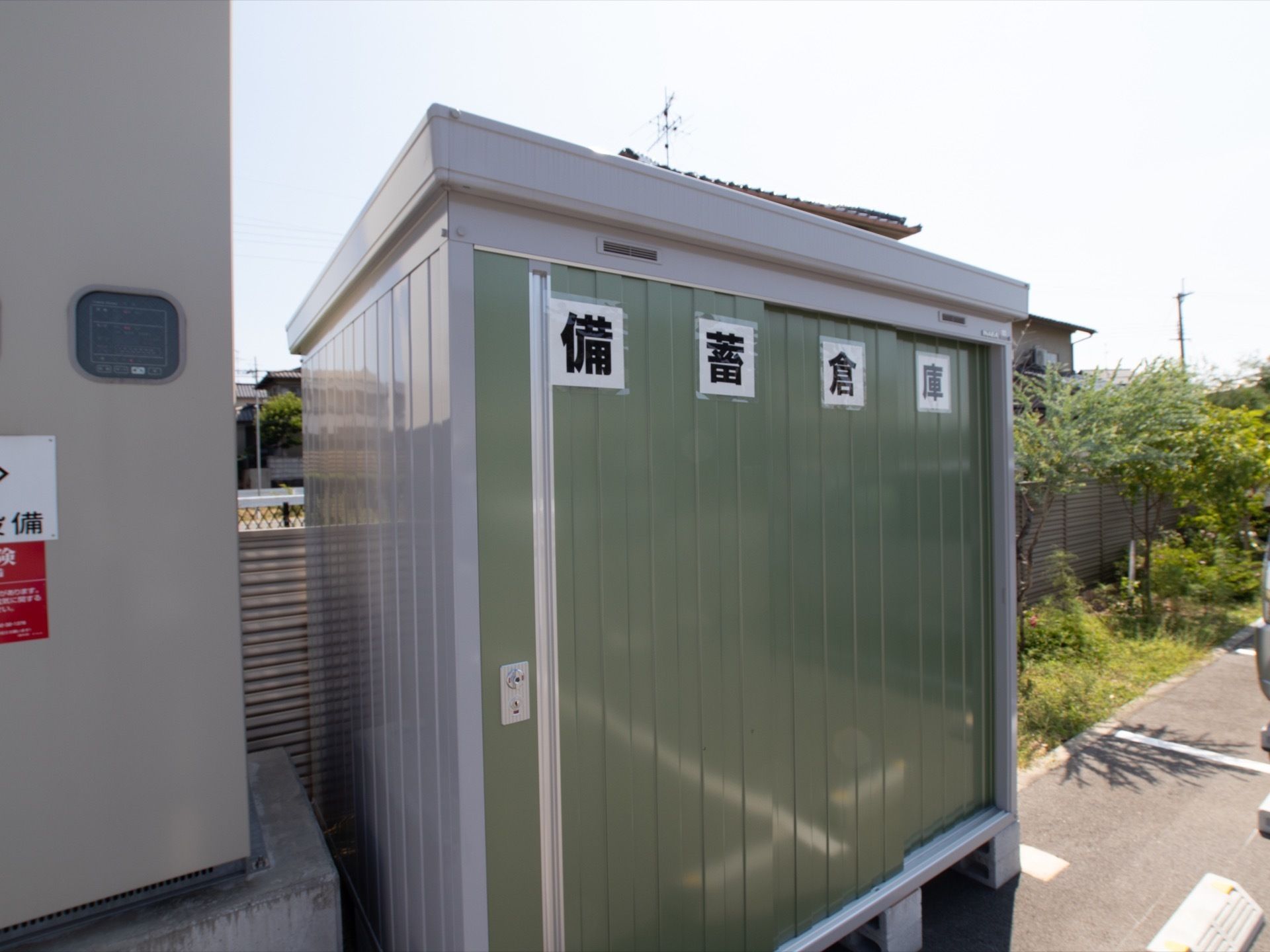 駐車場の一角に、備蓄品が保存されている倉庫がある。駐車場の周囲は芝生と木々で囲まれており、近隣は住宅街になっている。