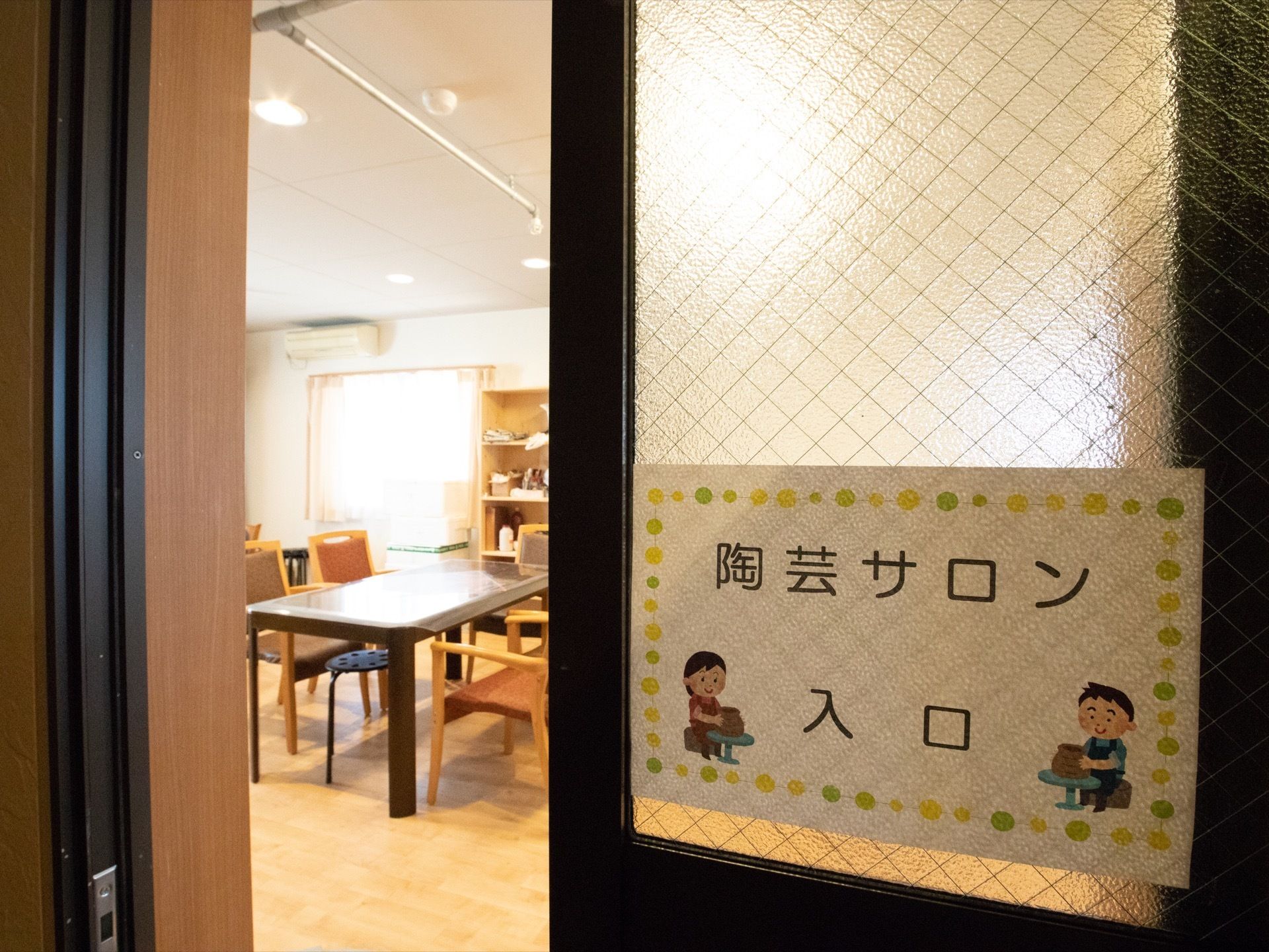 陶芸サロン入口と書かれた扉の先には、テーブルと椅子が置かれている。そのほか、棚やエアコンが設置されている。