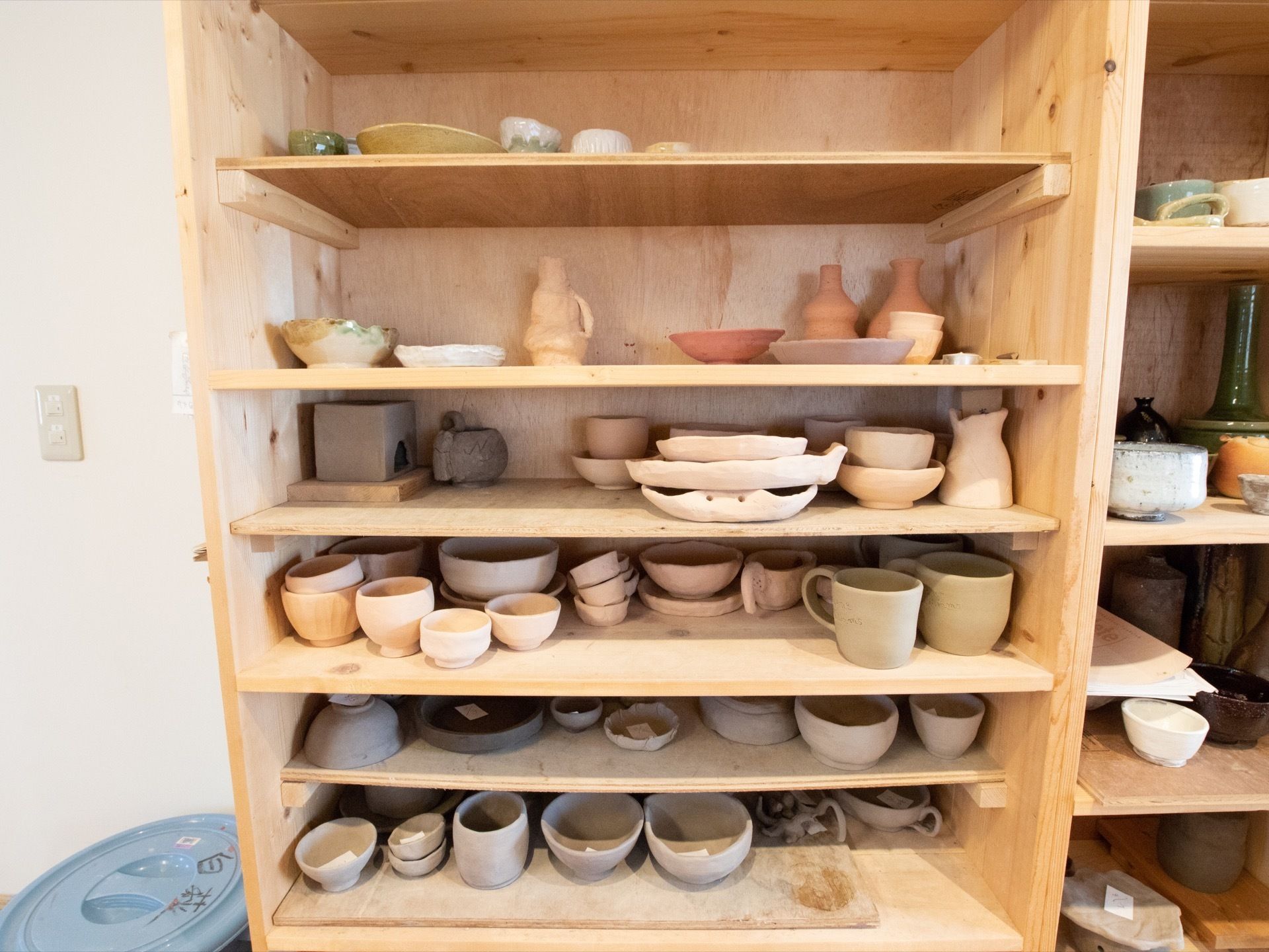 壁際に木製の大きな棚があり、お皿や水差し、お椀などの陶芸作品がたくさん並べられている。棚の脇にバケツ型のごみ箱がある。