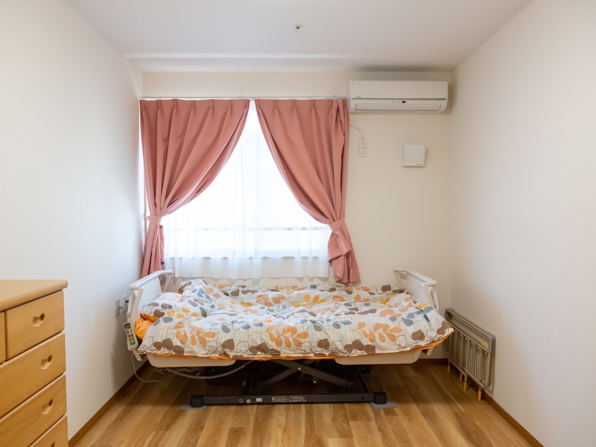 白い壁に木目調の床の居室である。介護用ベッドには、ポップな布団セットが敷いてある。エアコンを完備し、ピンク色のカーテンが掛かっている。