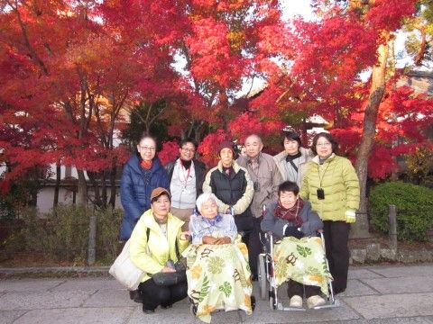 鮮やかな赤色に染まるモミジの木々の前で写真に収まるスタッフと入居者様