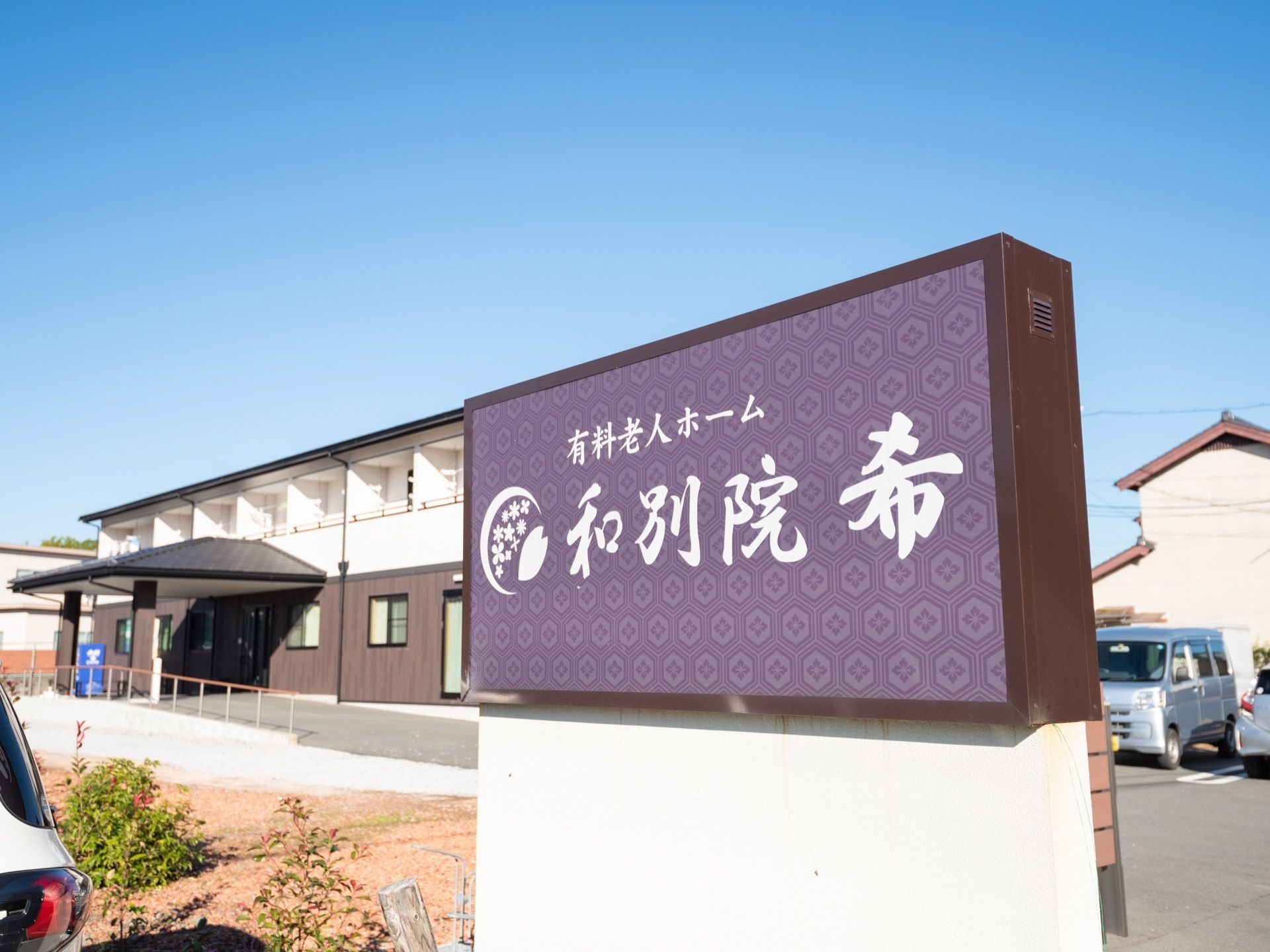施設敷地の前に設置されている施設看板である。紫色の和柄プレートに白いデザイン文字で施設名称と種別が明記されている。