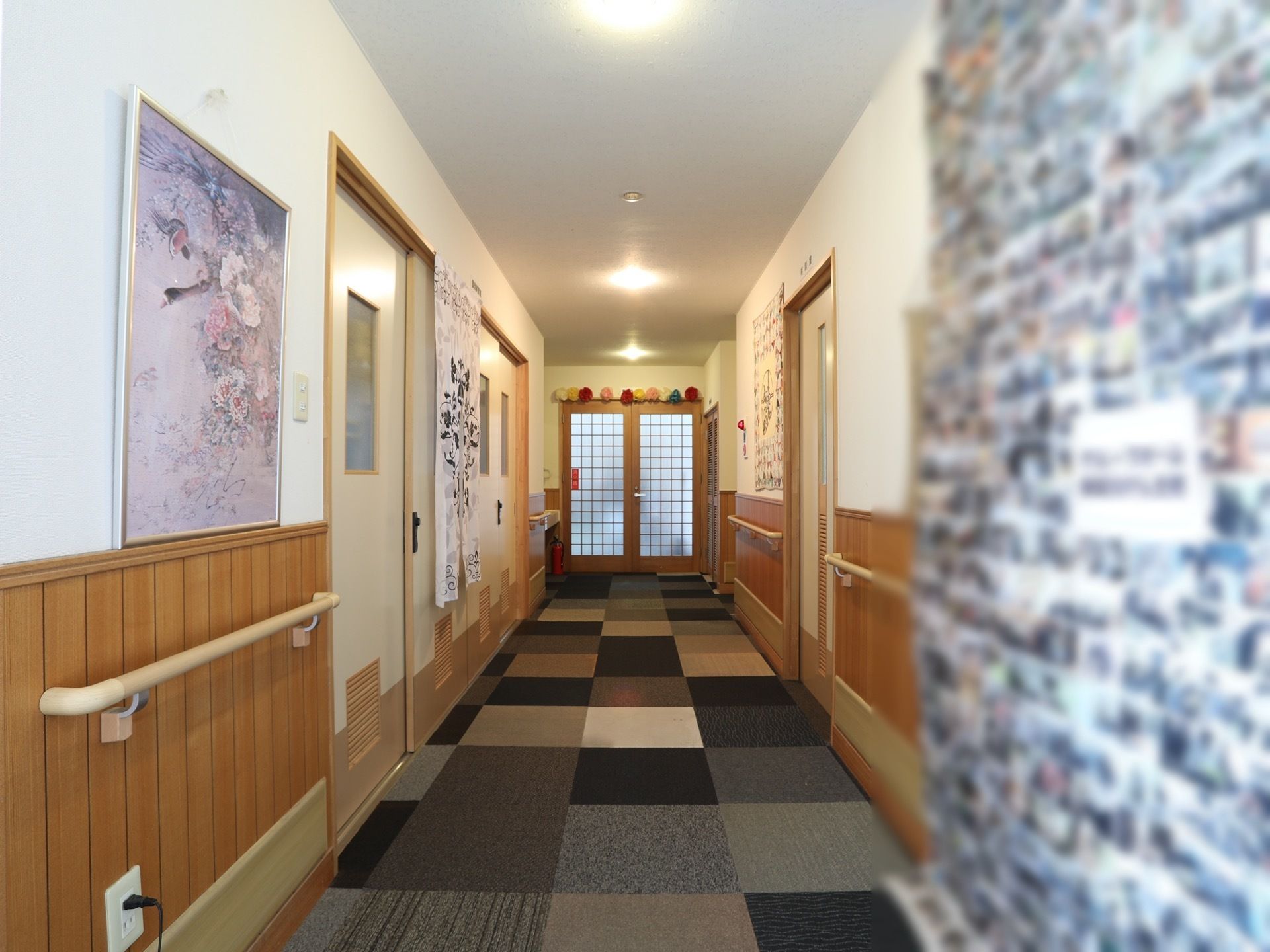 出入口へ続く通路は、絵画や入居者様の写真が飾られ親しみのある雰囲気である。左右の壁には、体を支えられるよう手すりが備え付けてある。