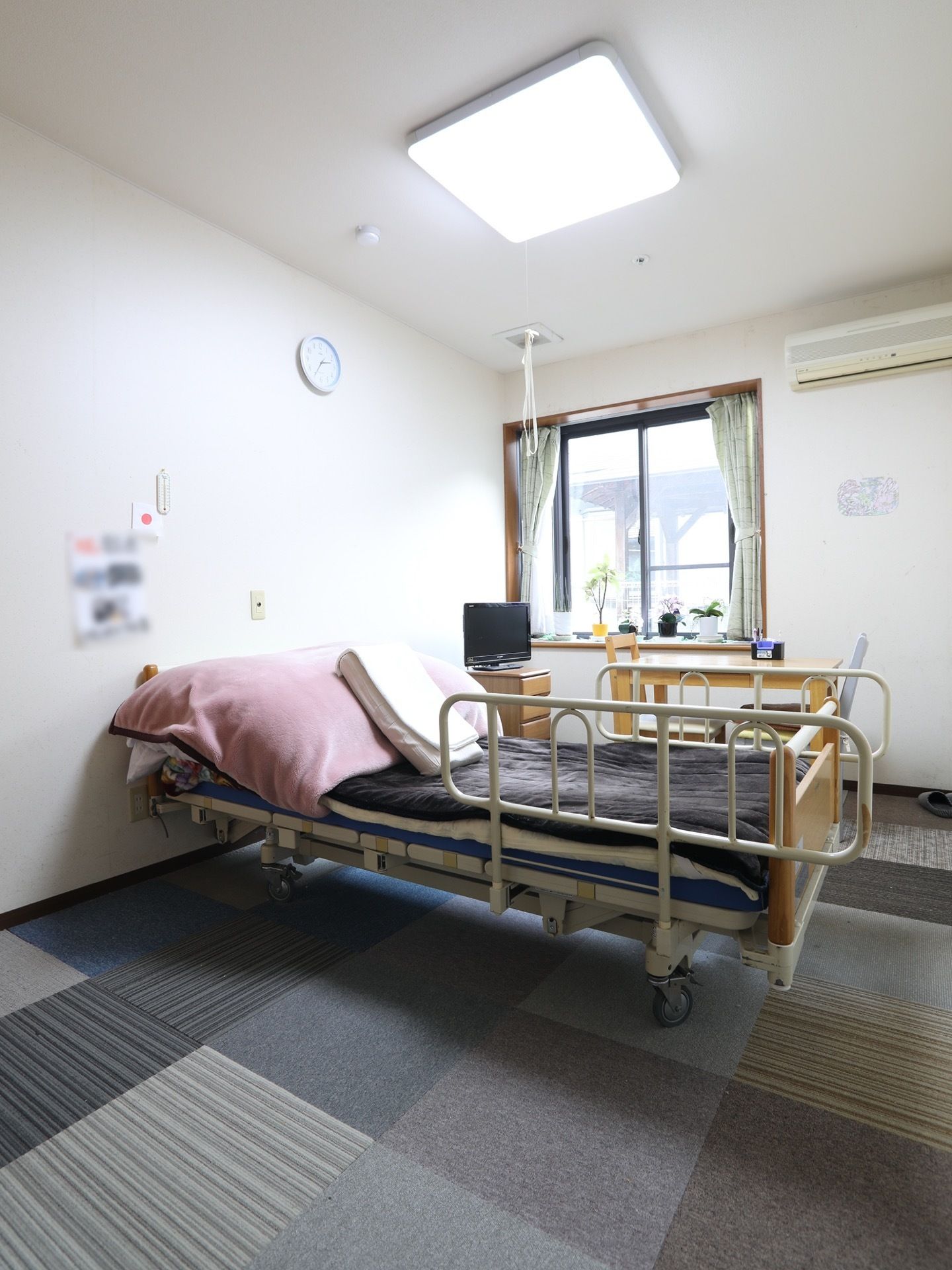 居室中央に介護ベッドとテーブルセットが置かれている。日当たりの良い窓があり、小さな観葉植物が並べられている。
