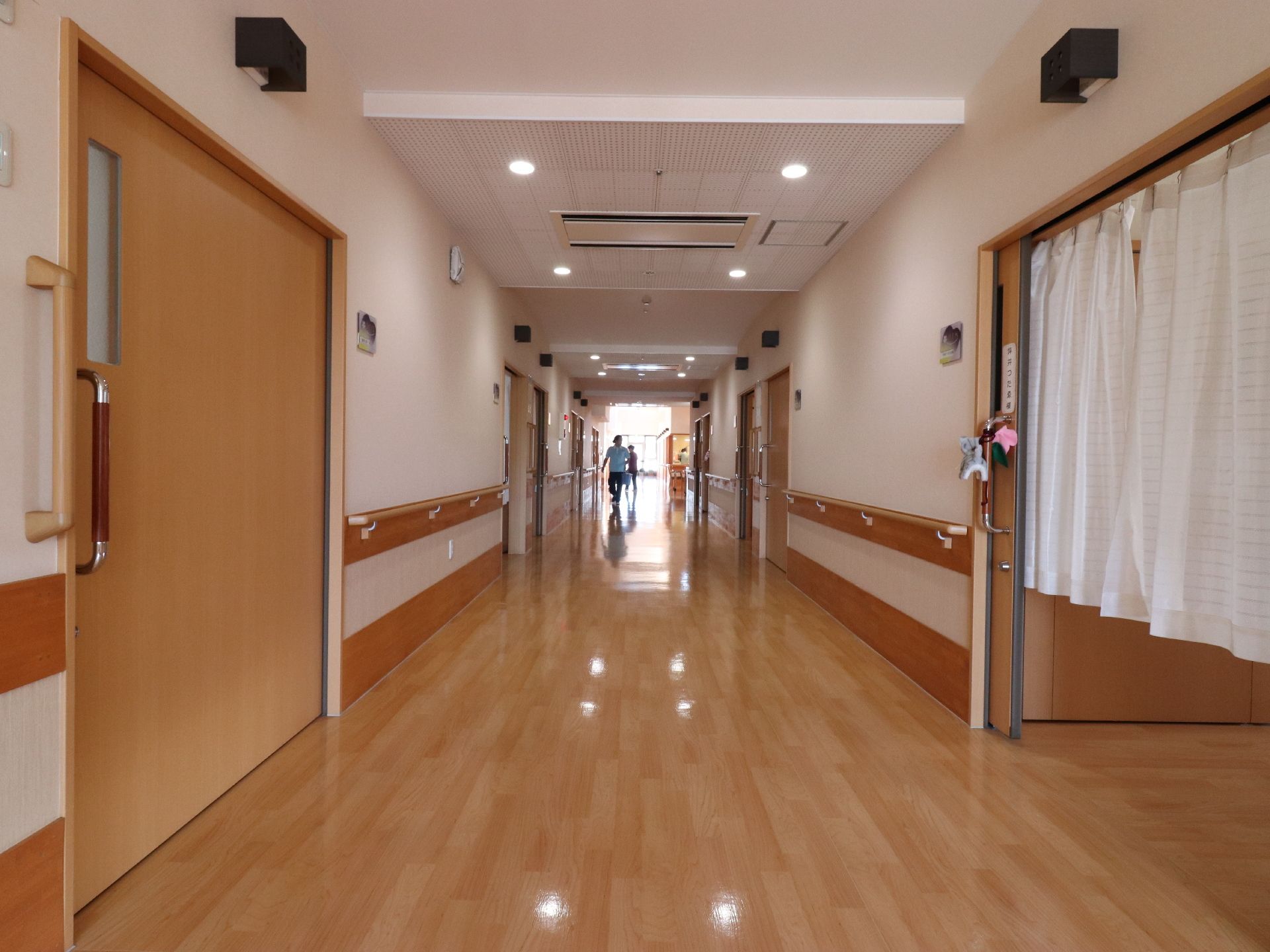 施設の廊下の様子。両側に居室のドアが並んでいる。廊下の先は窓になっており、明るい光が差している。奥に人が2人見える。