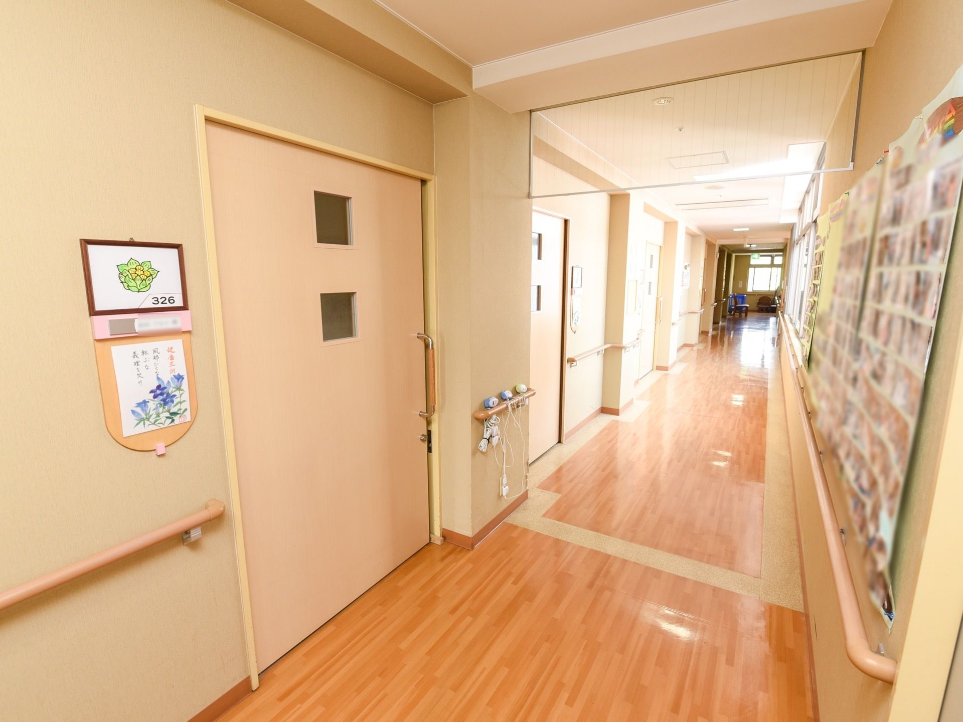 ベージュのドアが並ぶ、木目調の床の廊下である。壁に写真などを貼り、また、両側の壁には手すりを完備している。窓も設けられ明るい空間である。