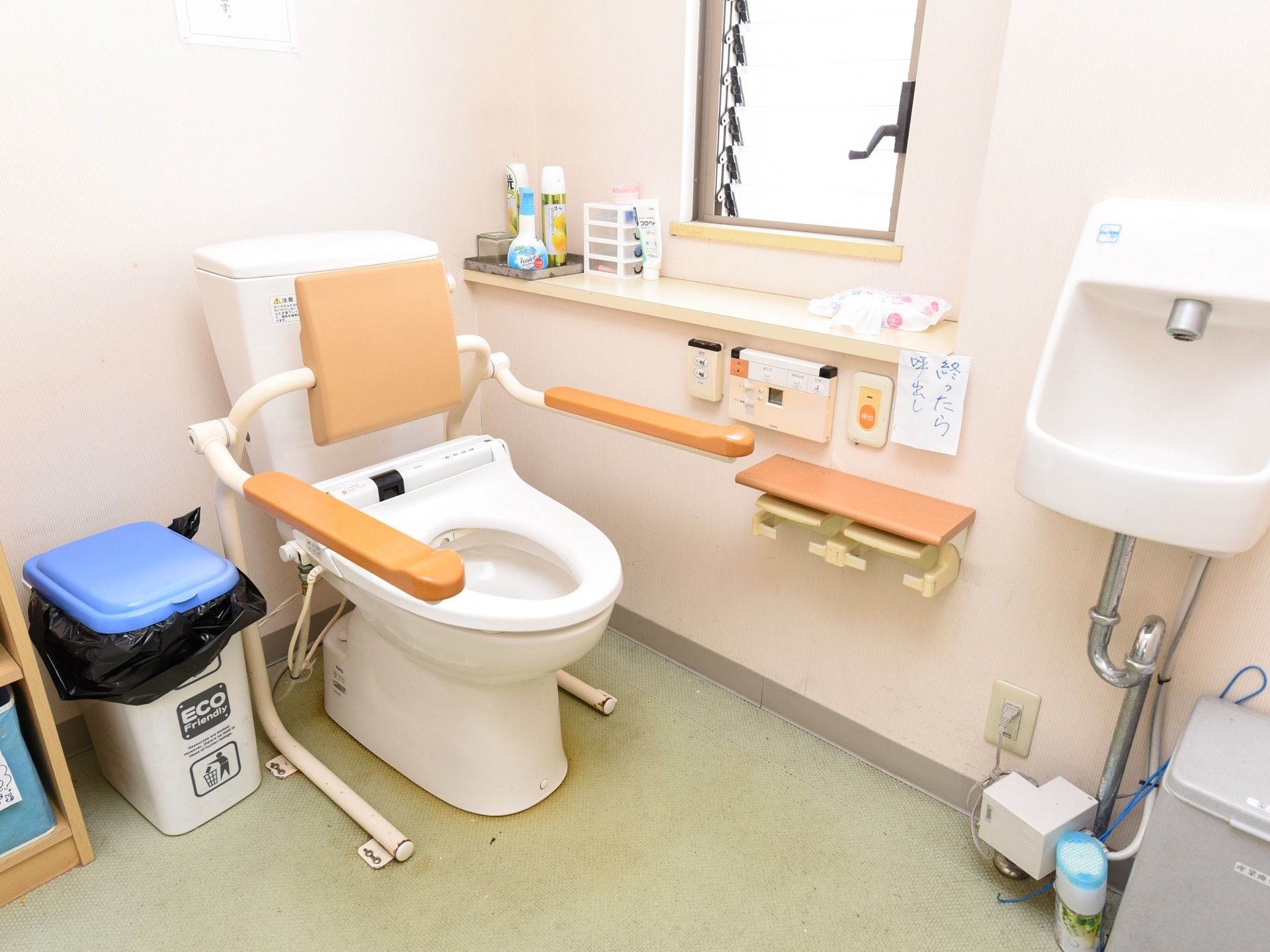 シンプルなデザインの洗面台を備えたトイレスペースである。便座には背もたれとひじ掛けを備えている。横側に窓と小物が置ける棚を設けている。