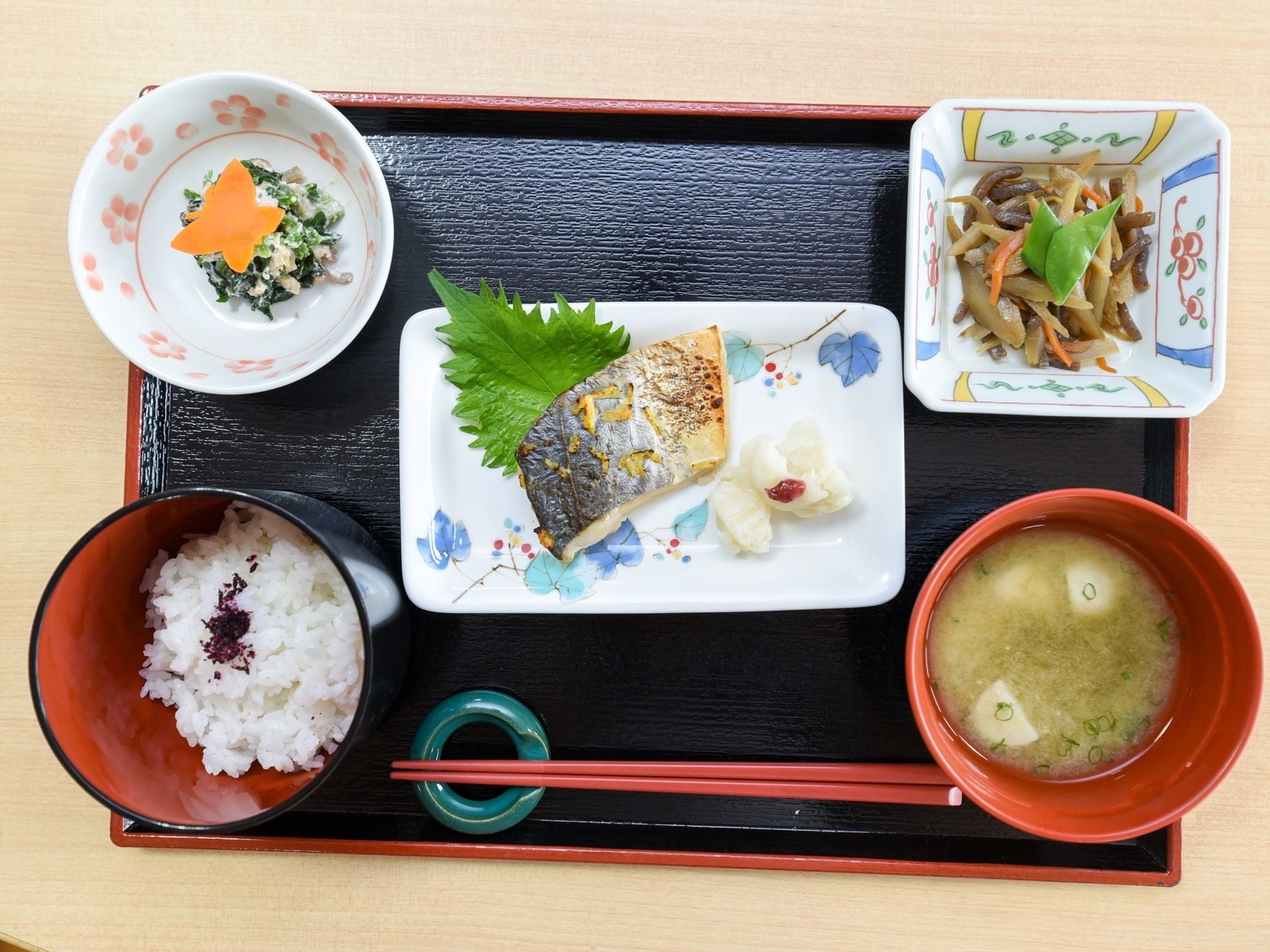 黒いトレーの中央にメインおかずの焼き魚が置かれ、手前にお箸、両側にご飯と味噌汁、上には２品の小鉢が添えられている。