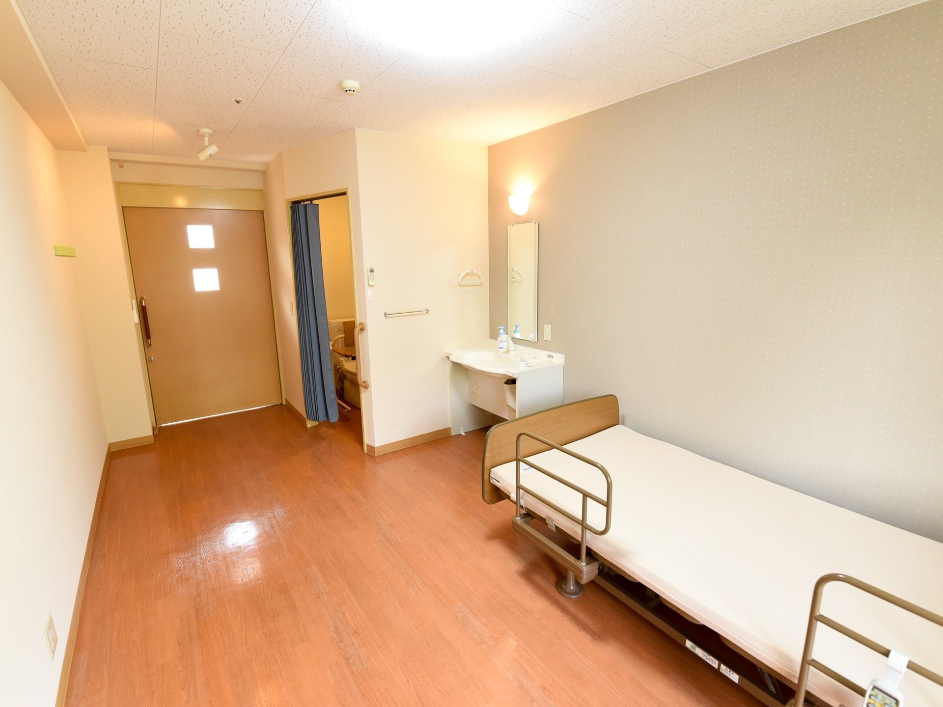 明るい木目調のフローリングの居室の壁際に、介護用ベッドを配置している。角に洗面台を設置し、出入口ドアの傍にトイレを設けている。