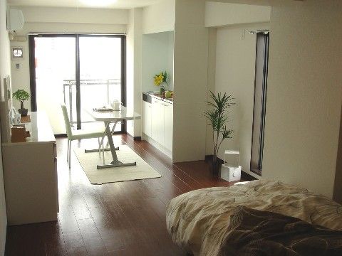奥の窓続きでベランダがあり、キッチンとテーブル、椅子、ベッドがある居住スペース