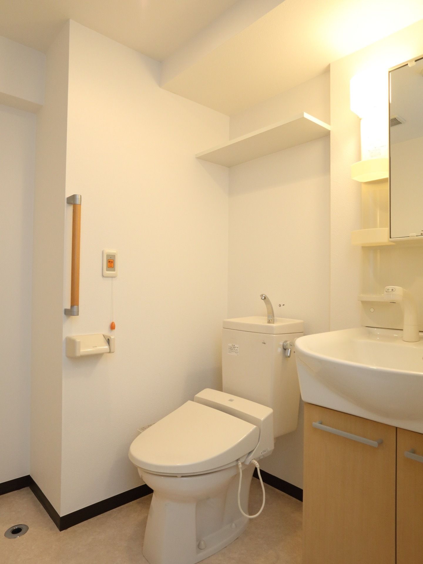 清潔感があふれるトイレの壁には手すりと緊急通報装置が設置されている。便器の上部には、棚が設けられている。